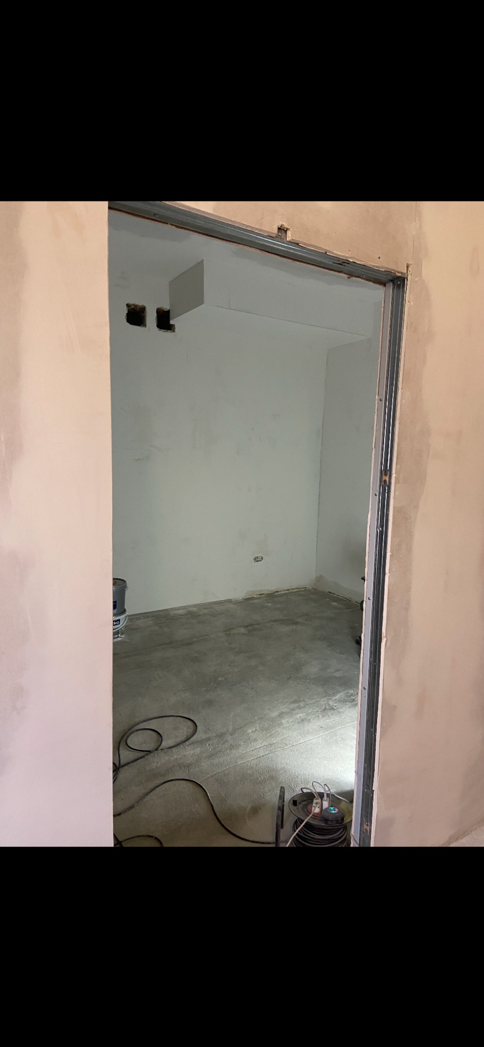 Wnętrze w trakcie remontu: surowy beton na podłodze, ściany częściowo pokryte gładzią, widoczne puszki elektryczne i rozciągnięty przewód zasilający.
