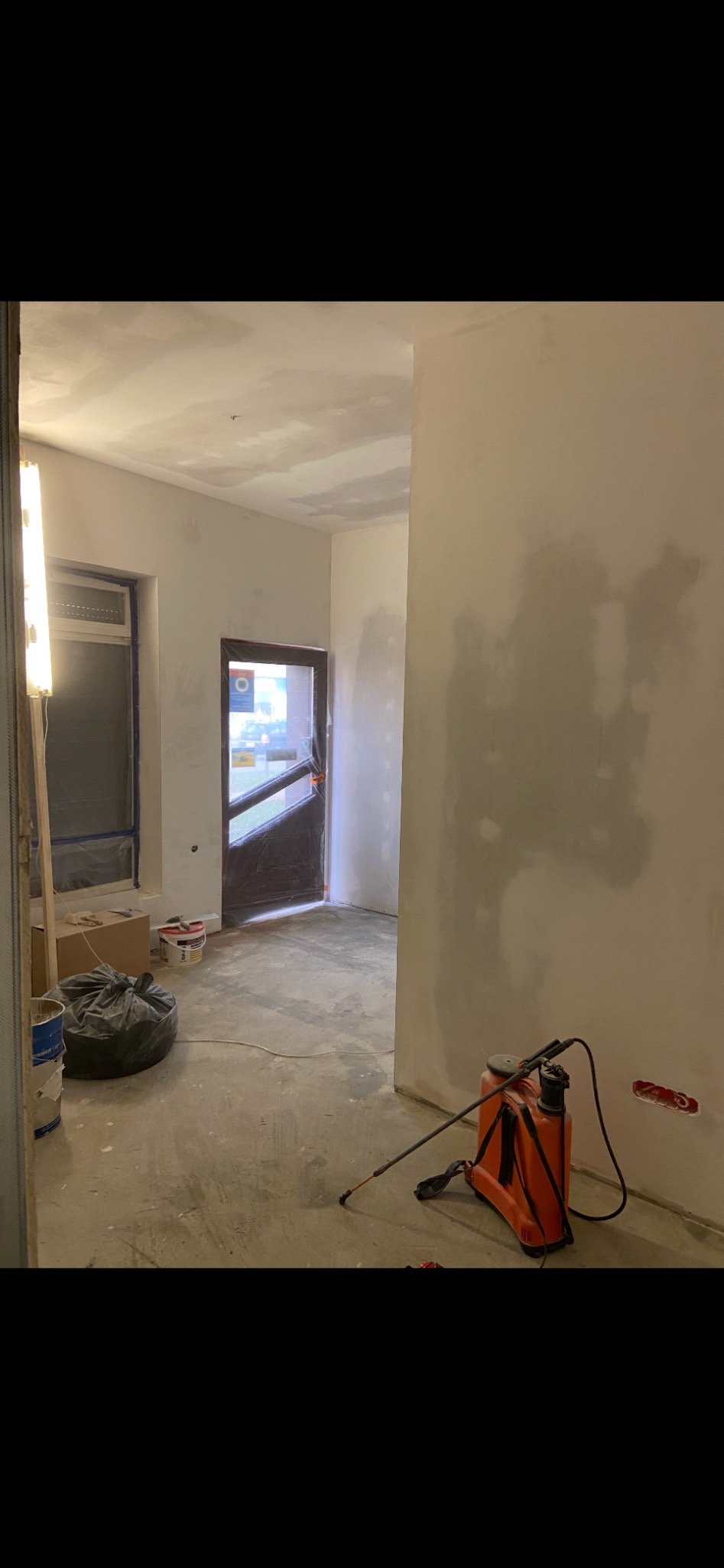 Pokój w trakcie prac wykończeniowych: ściany przygotowane pod malowanie, częściowo zagruntowane, widoczny agregat malarski, okno zabezpieczone folią, surowa podłoga.