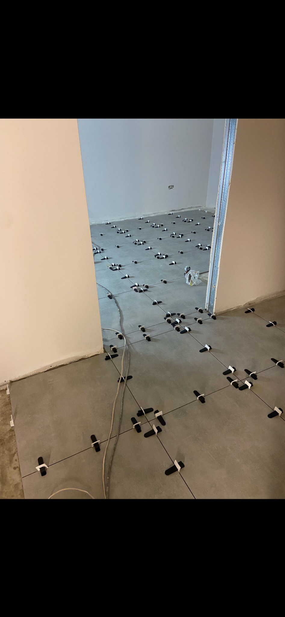 Układanie szarych płytek podłogowych z czarnymi krzyżykami dystansowymi w trakcie remontu pomieszczenia, widoczne kable elektryczne na podłodze.