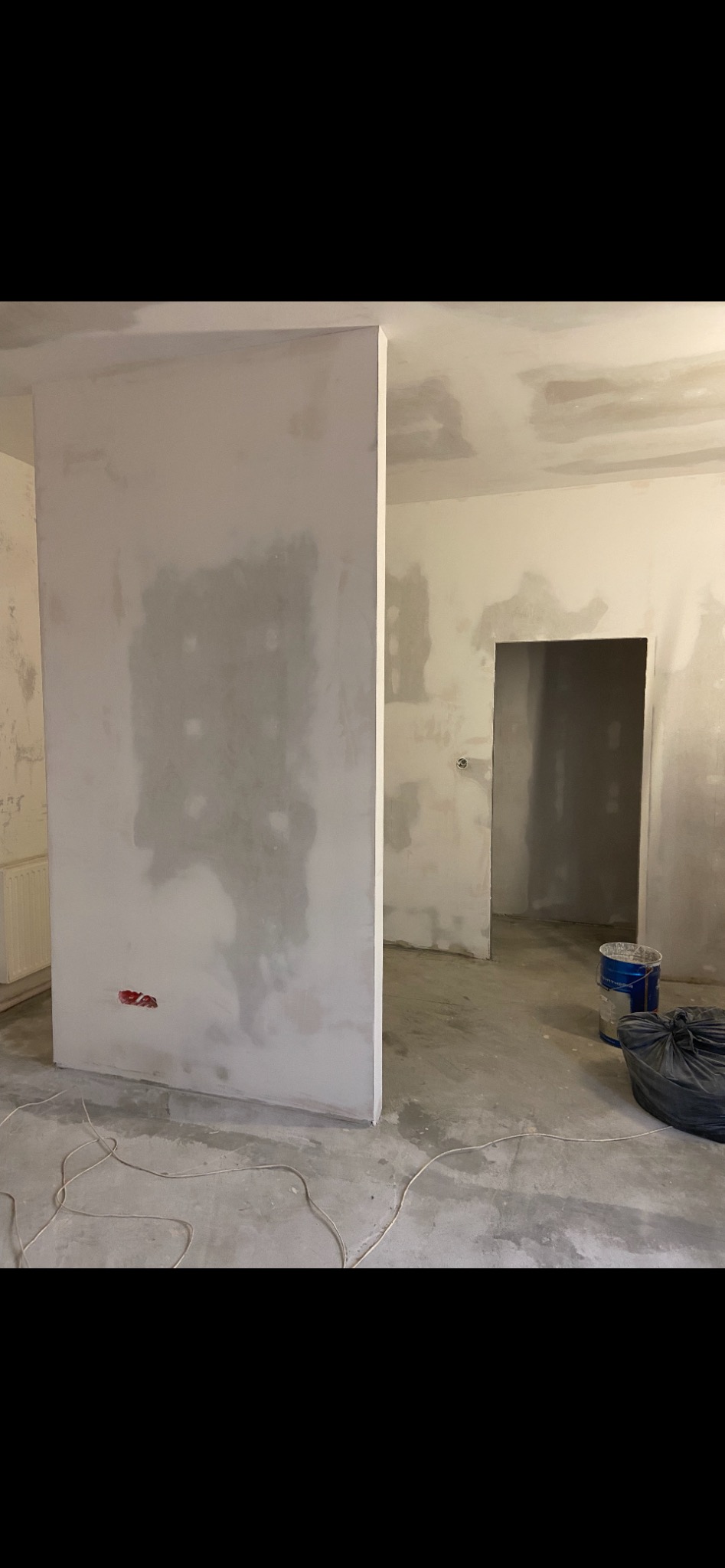 Wnętrze w trakcie remontu: ściana z płyt kartonowo-gipsowych przed szpachlowaniem, widoczne puszki elektryczne, surowy beton na podłodze, wiadro z farbą i worek na śmieci w tle.