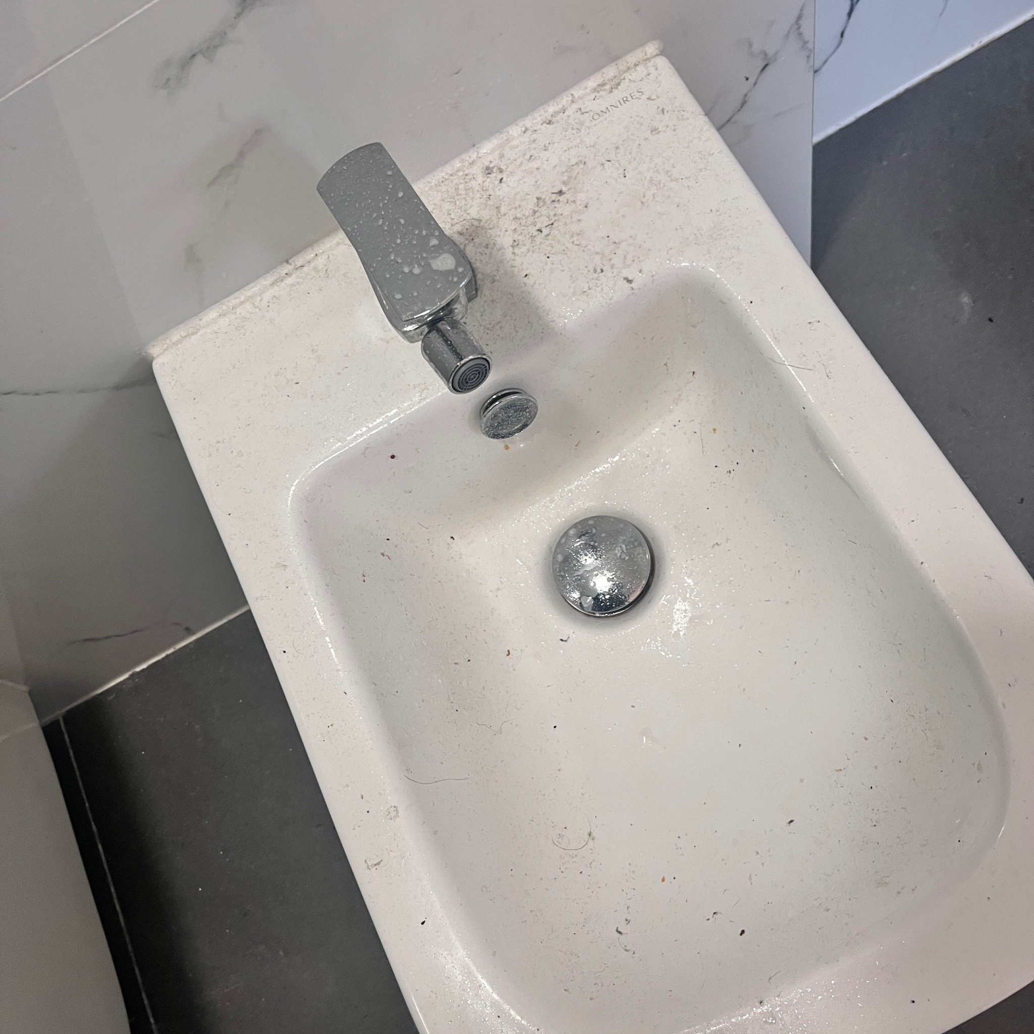 Brudny bidet z chromowaną baterią i odpływem, widoczne zabrudzenia na ceramice i armaturze, tło stanowi marmurowa okładzina ścienna i ciemna podłoga.