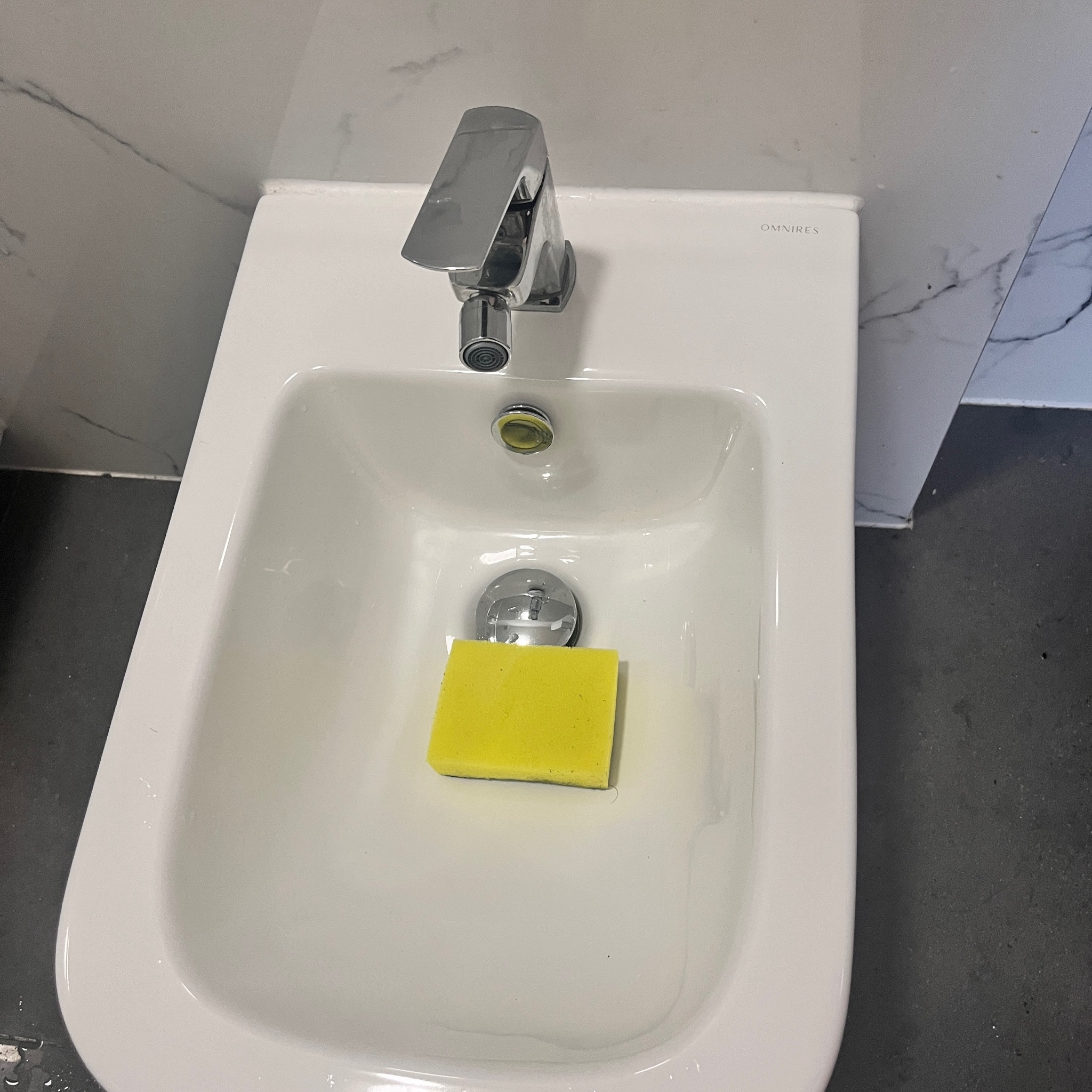 Bidet marki Omnires z chromowaną baterią i żółtą gąbką w środku, na tle marmurowej ściany i ciemnej podłogi.