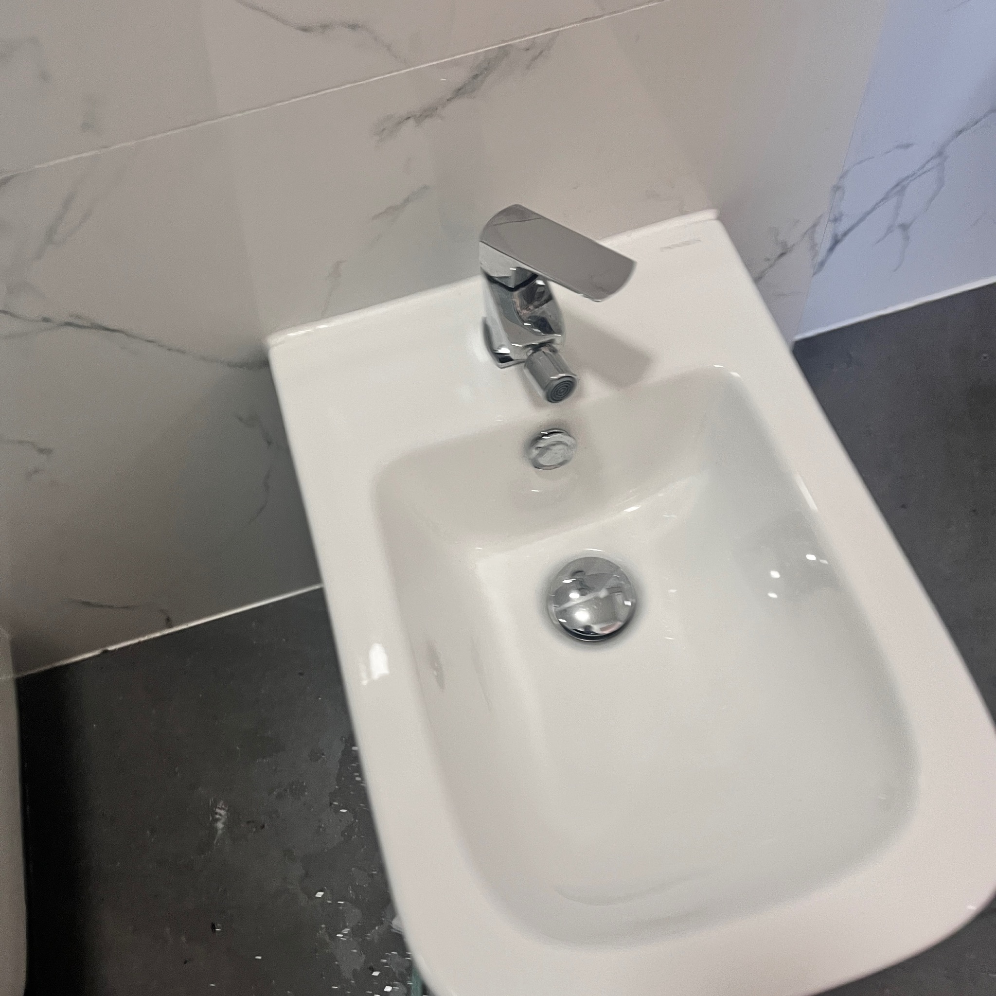 Bidet z nowoczesną, chromowaną baterią, zamontowany przy ścianie wyłożonej marmurowymi płytkami. Widoczne szare fugi i ciemna podłoga z drobnymi plamami wody.