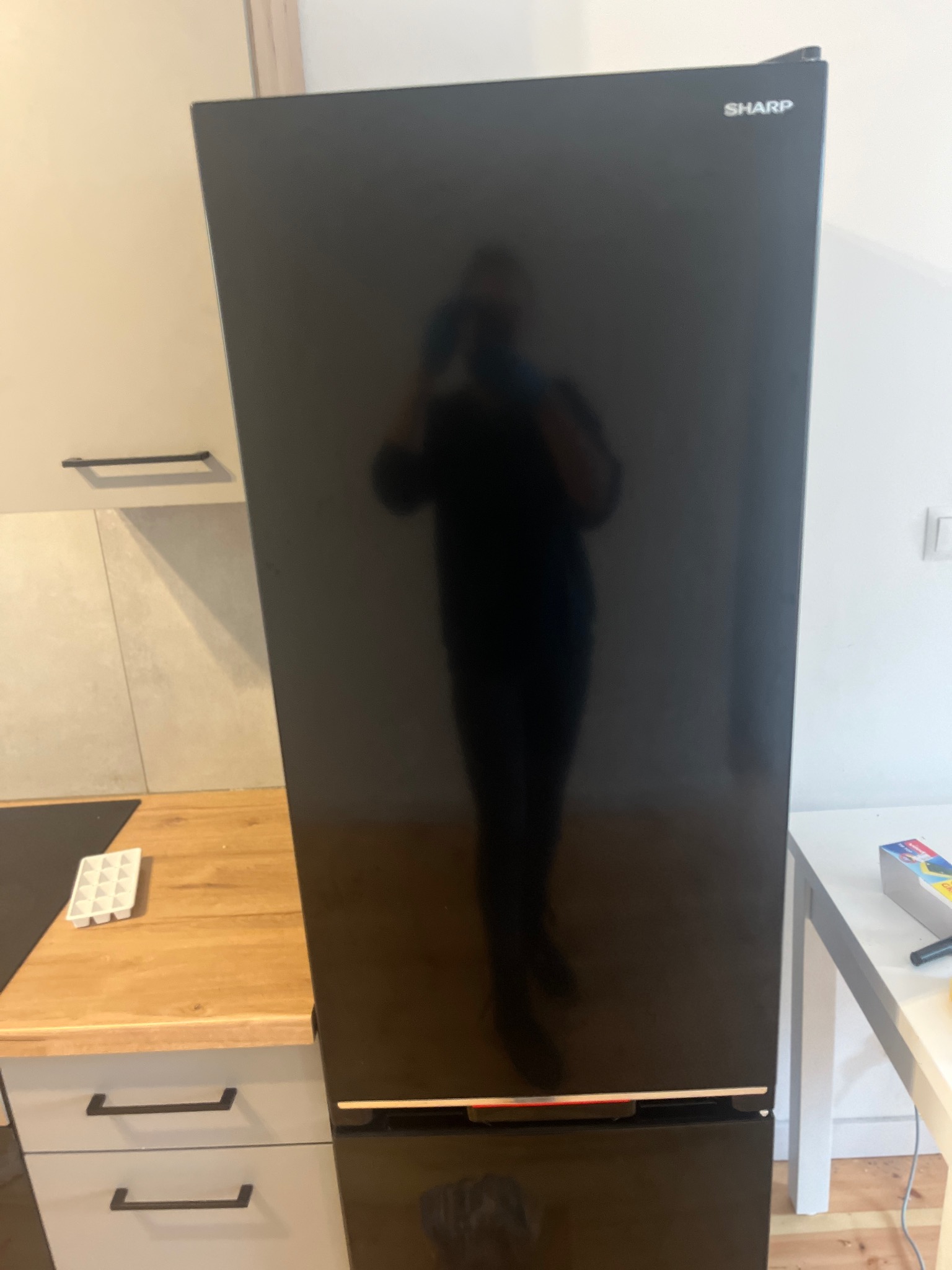 Czarna lodówka marki Sharp w kuchni z blatem drewnopodobnym i białymi szafkami, widoczne odbicie osoby robiącej zdjęcie.