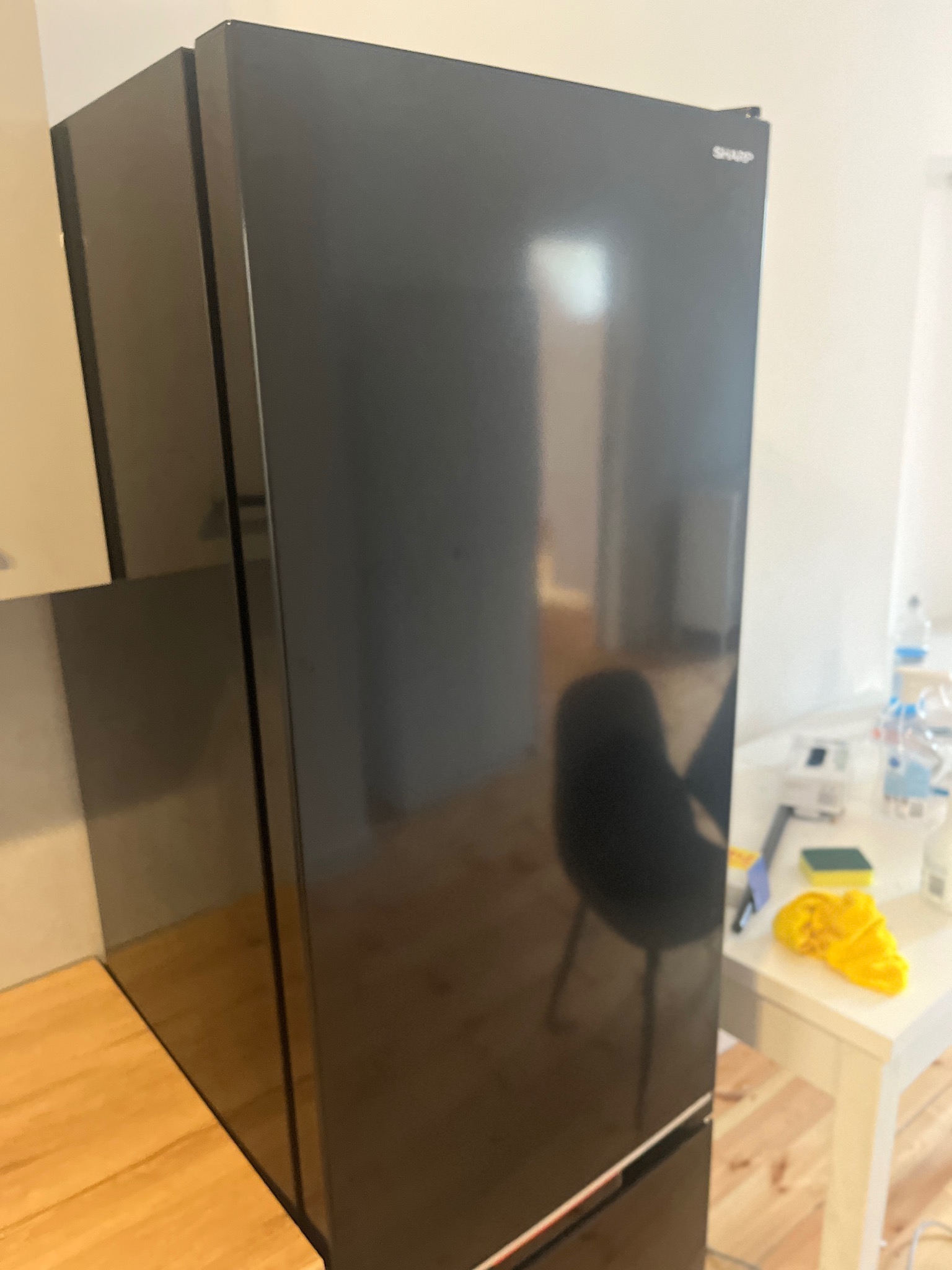 Błyszcząca, czarna lodówka marki Sharp stoi w kuchni; na blacie obok widoczne akcesoria do sprzątania, w tym żółta ścierka i gąbka.