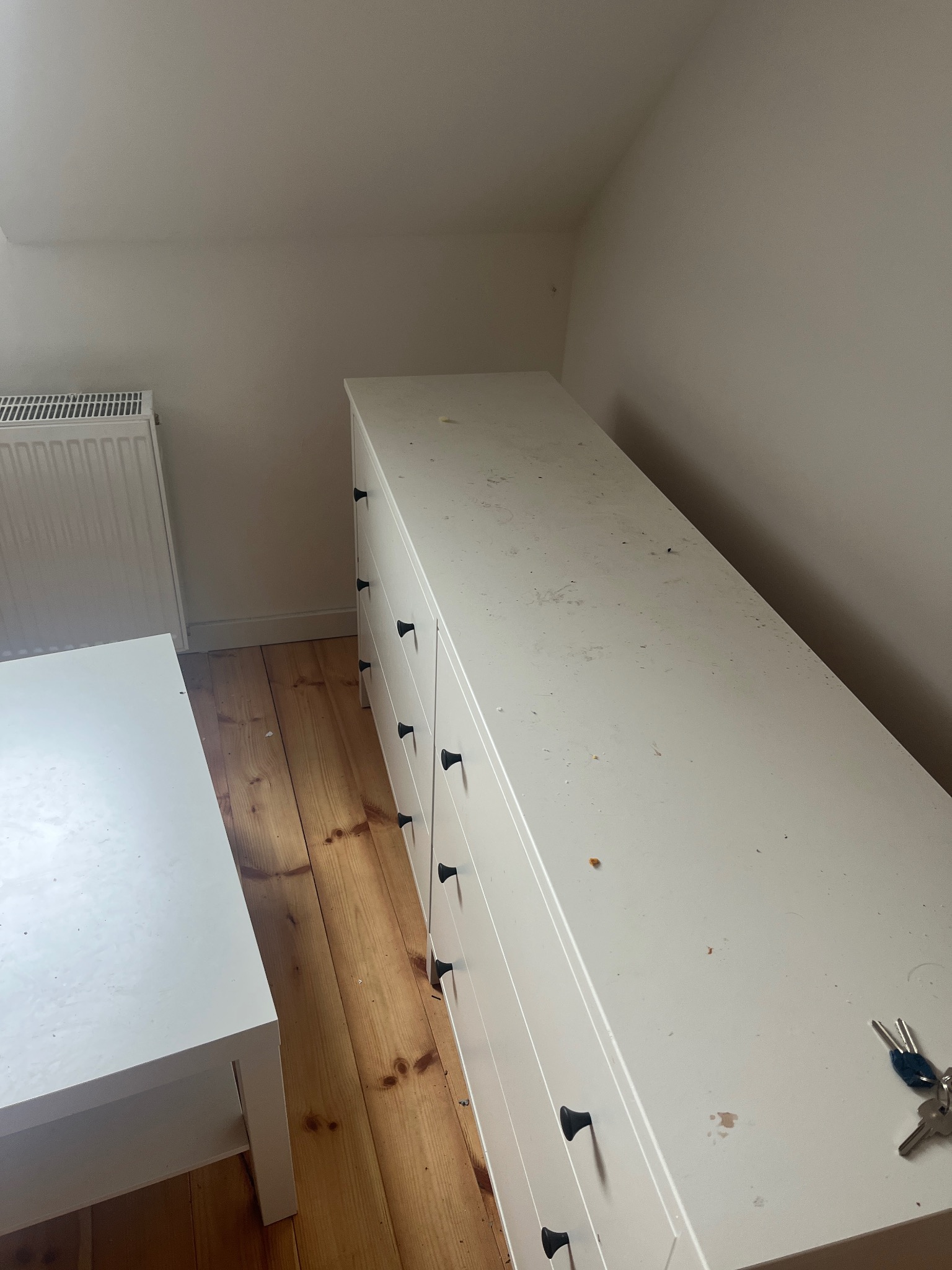 Pokój na poddaszu przed wysprzątaniem: biała komoda z ciemnymi uchwytami, drewniana podłoga, biały stolik i grzejnik. Widoczne zabrudzenia i klucze na komodzie.