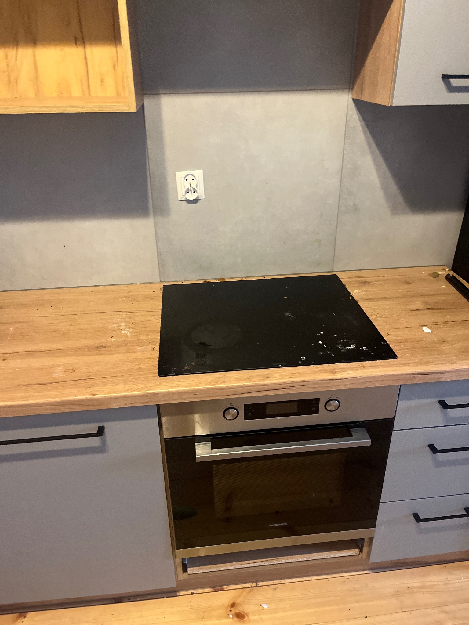 Fragment kuchni po montażu płyty indukcyjnej i piekarnika, widoczne ślady po remoncie na blacie, szare szafki z czarnymi uchwytami i kontakt elektryczny na ścianie.
