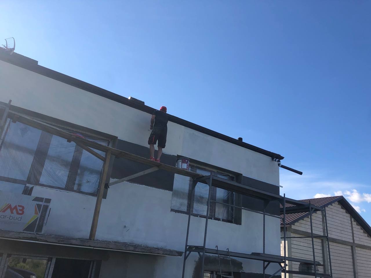 Mężczyzna w czerwonej czapce maluje elewację domu z rusztowania; okna zabezpieczone folią, widoczna puszka z farbą.
