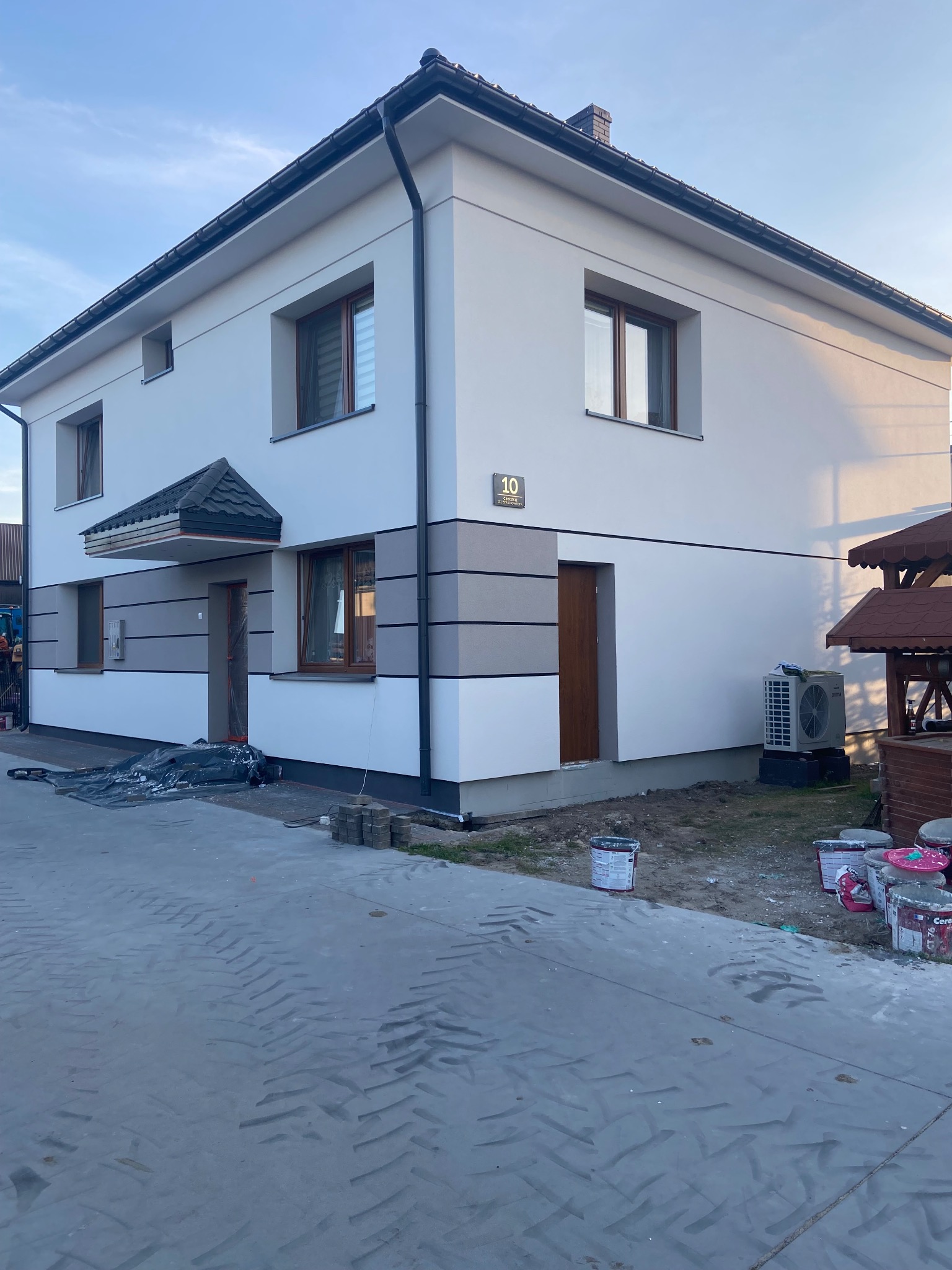Dwukondygnacyjny dom z nową elewacją w odcieniach bieli i szarości, widoczne ślady prac malarskich, puste puszki po farbie i folia zabezpieczająca.