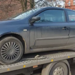 Brudny, granatowy samochód osobowy na platformie lawety, widoczny budynek z czerwonej cegły w tle.