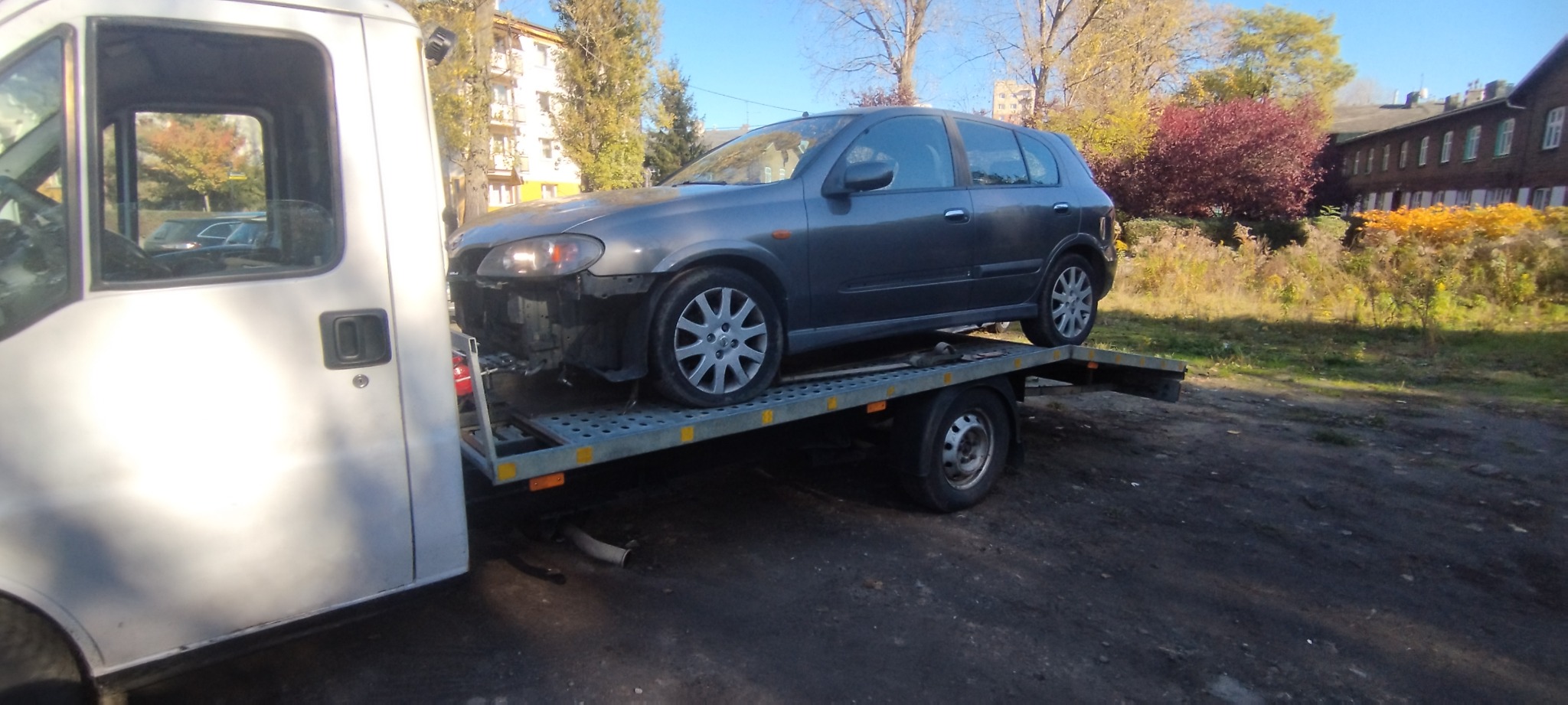 Uszkodzony, szary samochód osobowy bez przedniego zderzaka transportowany na lawecie w słoneczny dzień. W tle budynek mieszkalny i drzewa.