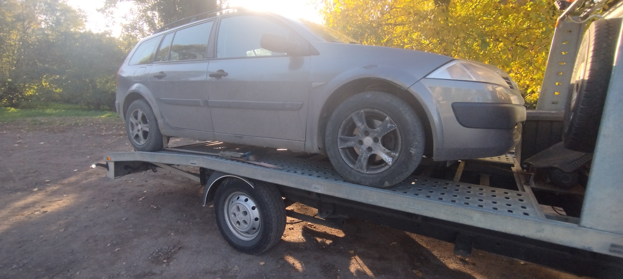 Szary samochód kombi marki Renault na ażurowej platformie lawety, widoczne drzewa w tle, oświetlone popołudniowym słońcem.