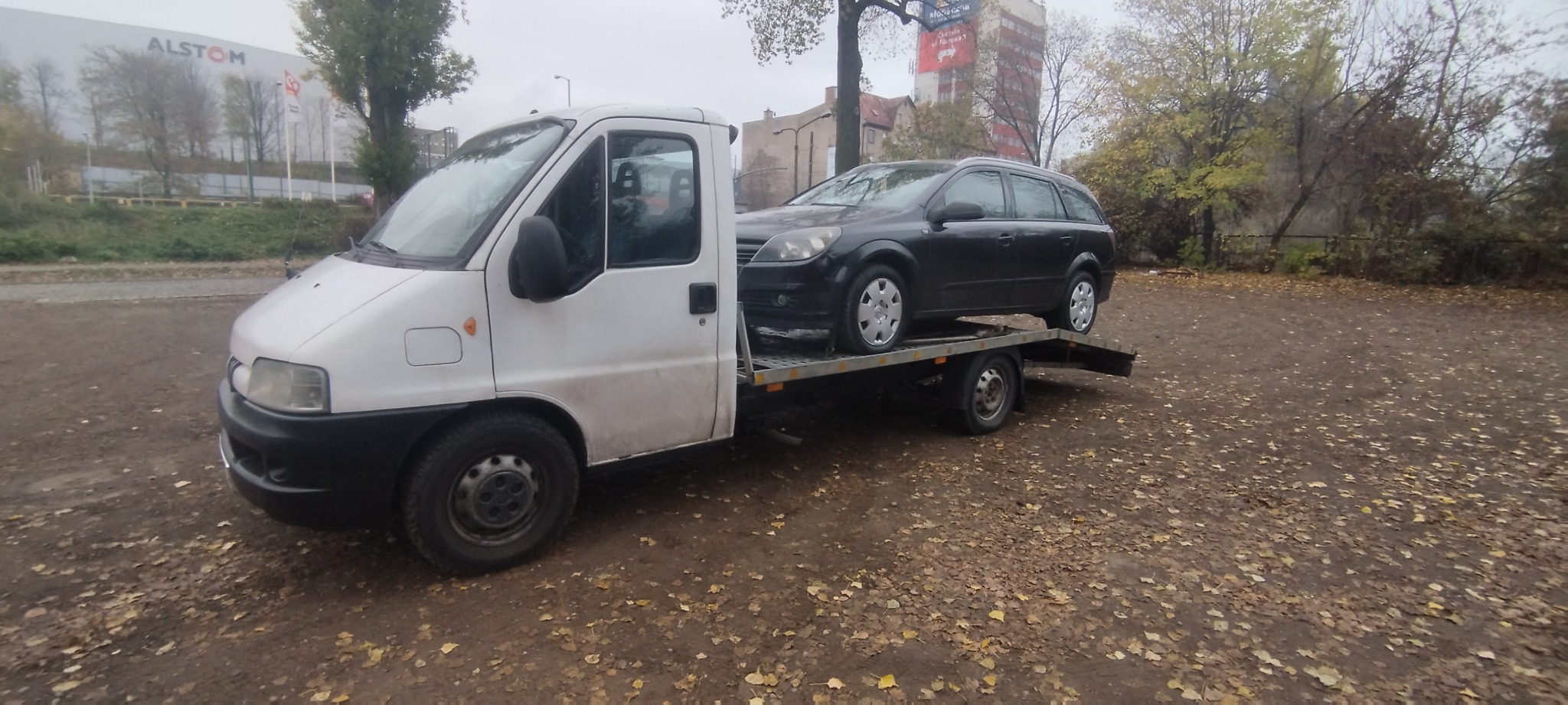Biała laweta z czarnym samochodem osobowym na platformie, zaparkowana na placu z opadłymi liśćmi, w tle budynki i drzewa w pochmurny dzień.