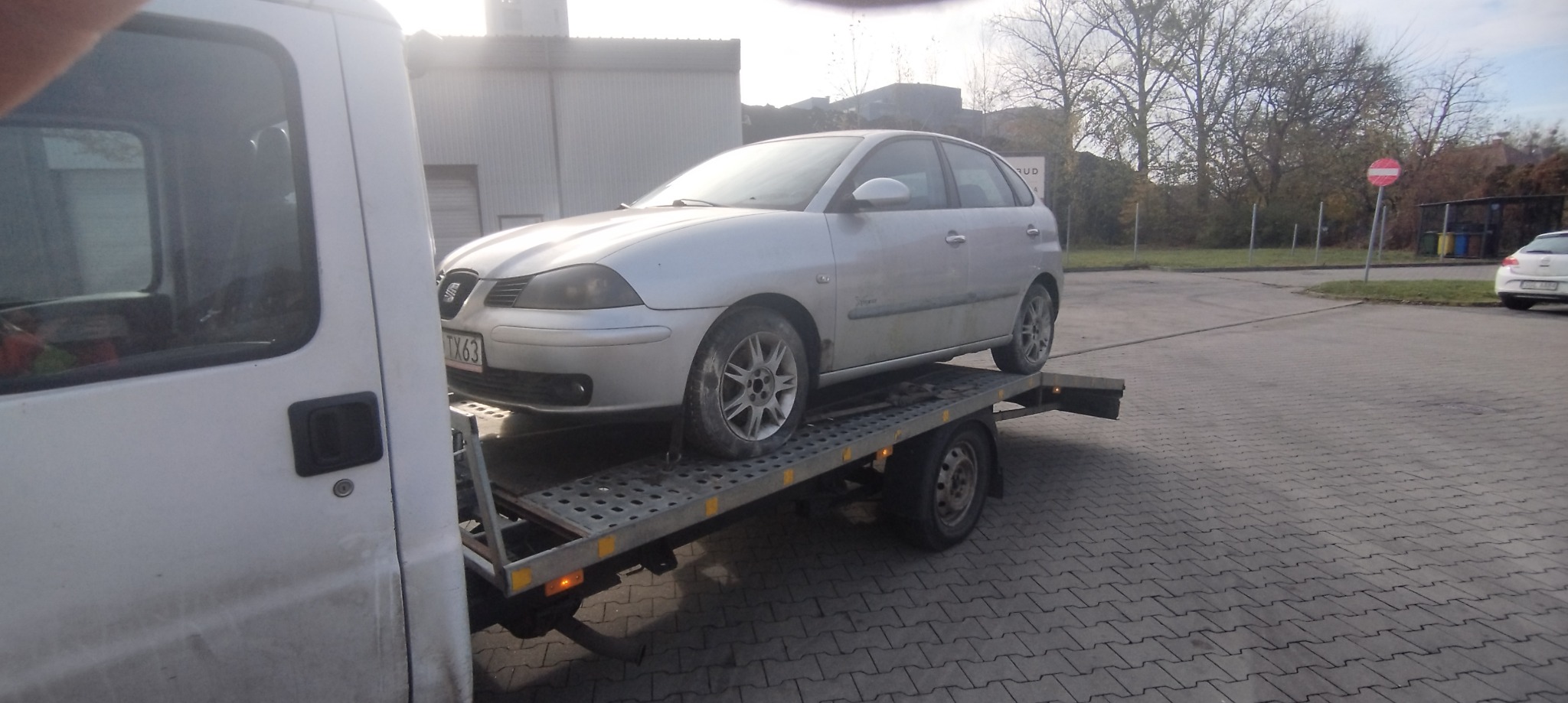 Srebrny samochód Seat Ibiza z polskimi tablicami rejestracyjnymi TX63 przewożony na metalowej platformie lawety, z widocznym fragmentem białego pojazdu holowniczego i otoczeniem placu z kostki...