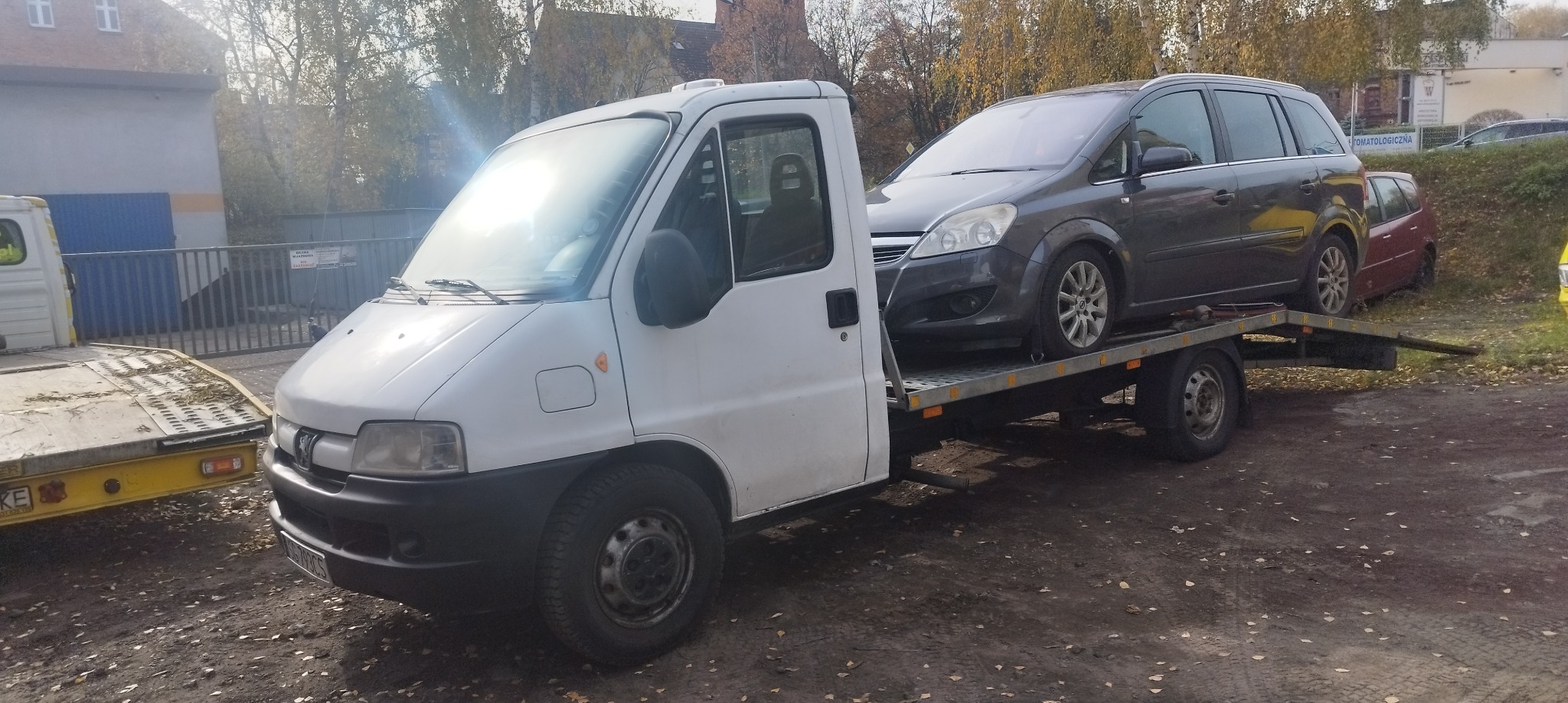 Biała laweta Peugeot z szarym samochodem osobowym Opel Zafira na platformie transportowej, zaparkowana na placu.