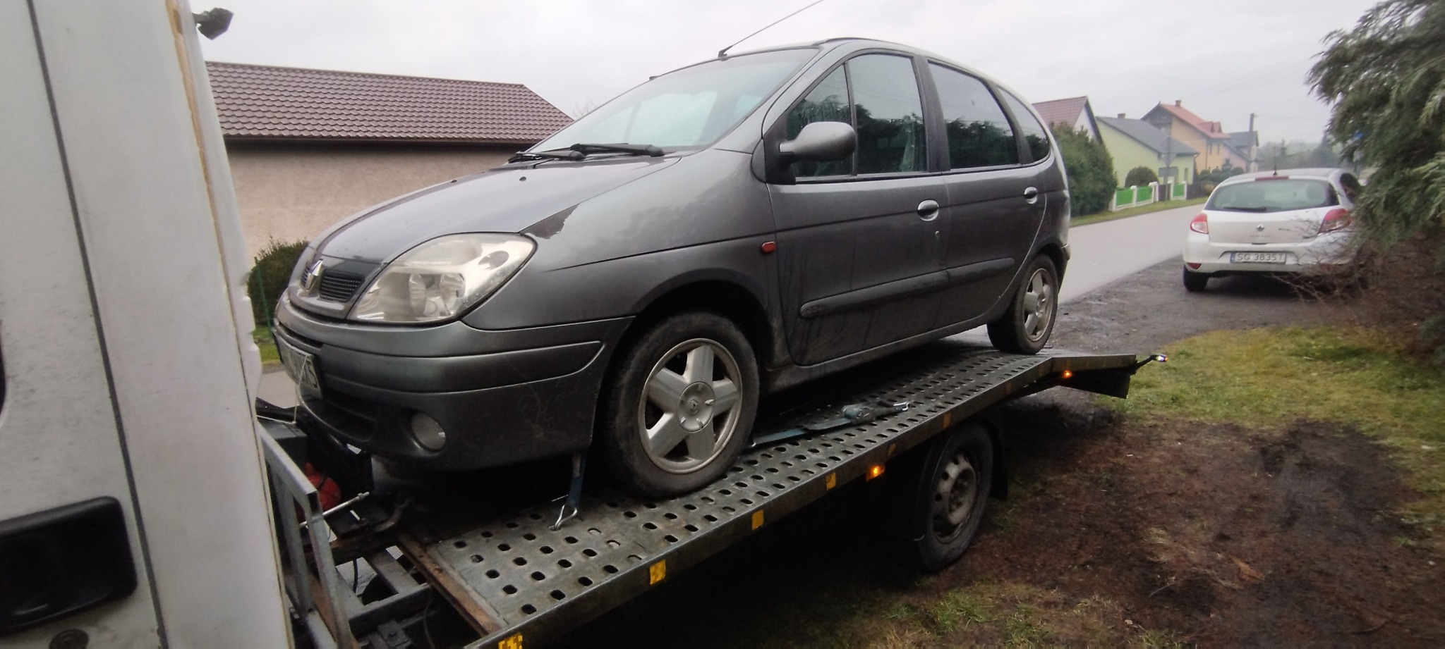 Szary samochód Renault Scenic zabezpieczony pasami transportowymi na ażurowej platformie lawety, w tle zabudowa jednorodzinna i biały samochód osobowy.