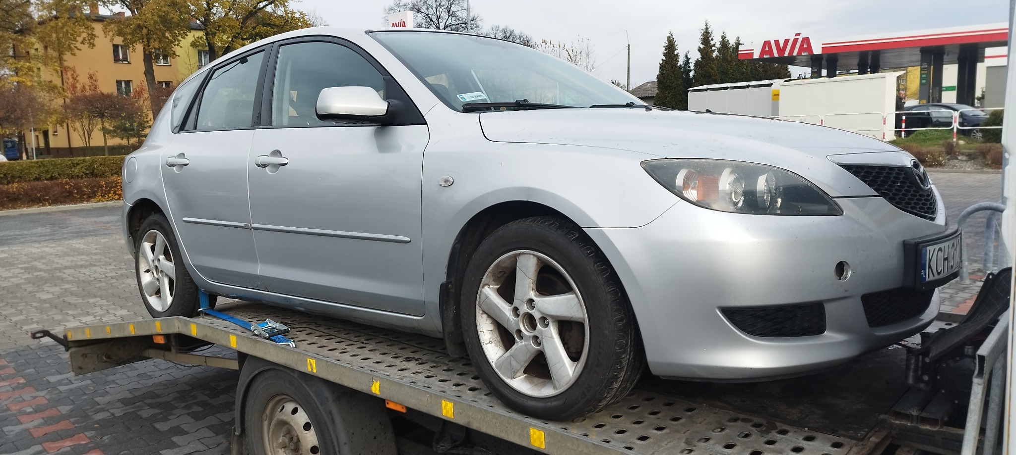Srebrny hatchback Mazda 3 zabezpieczony pasami na platformie lawety, widoczna stacja benzynowa Avia w tle.