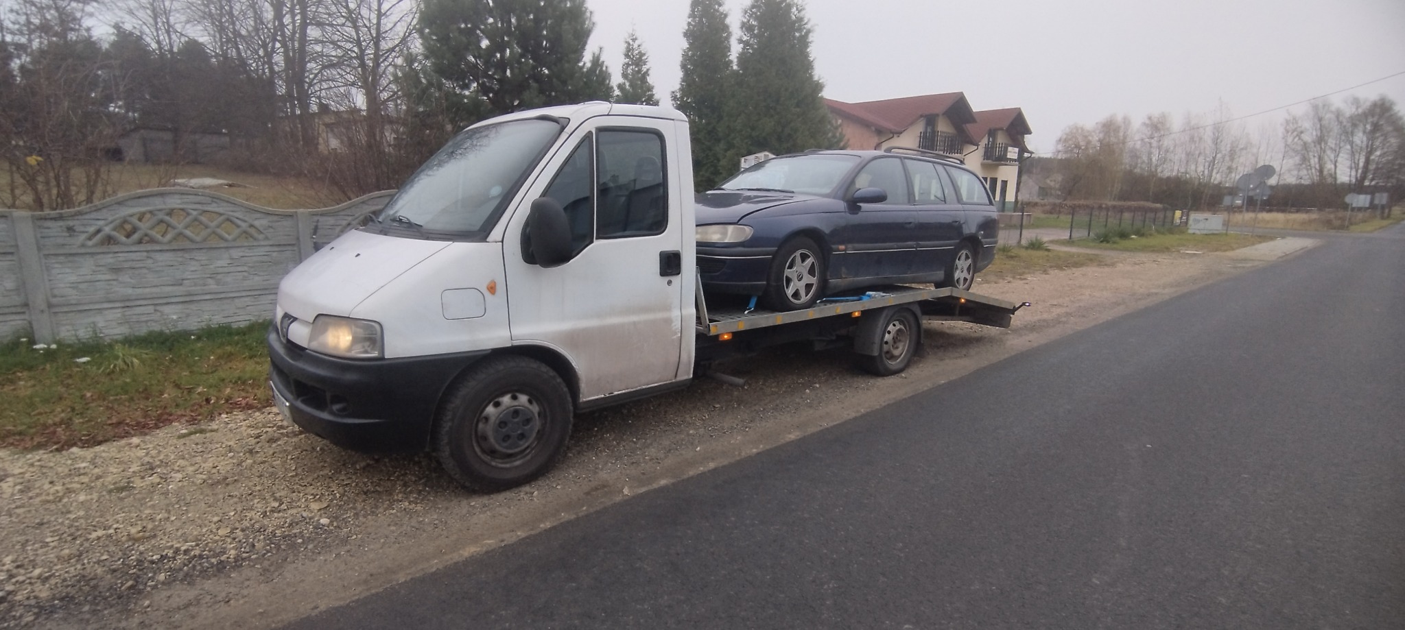 Biała laweta z niebieskim samochodem osobowym na platformie, zaparkowana na poboczu drogi w pochmurny dzień, widoczny fragment betonowego ogrodzenia i dom w tle.