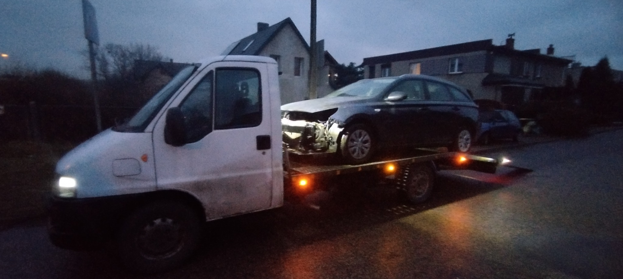 Uszkodzony, ciemny granatowy kombi na platformie lawety, transportowany wieczorem na tle zabudowań mieszkalnych.