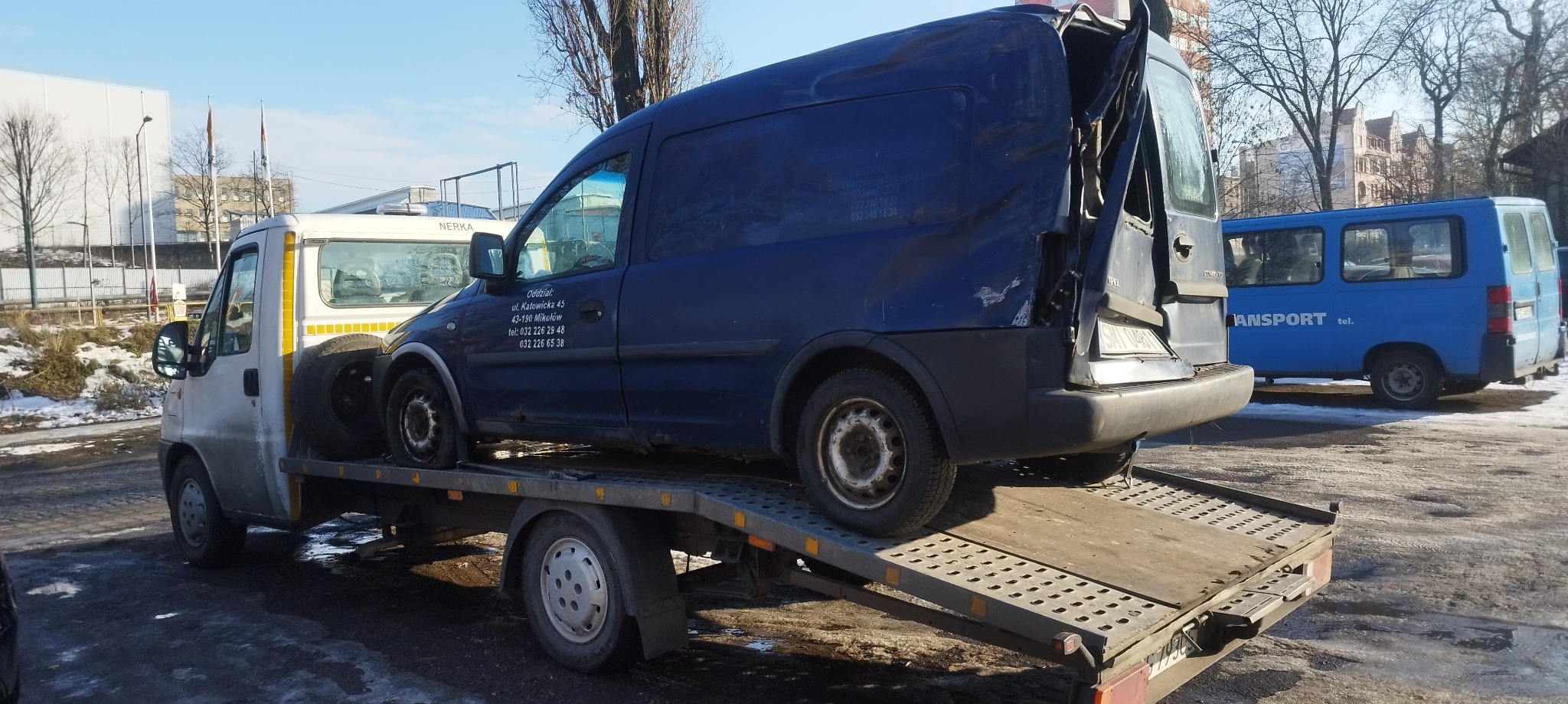 Uszkodzony, granatowy Opel Combo z otwartymi drzwiami, przewożony na lawecie w zimowych warunkach miejskich, z widocznymi śladami korozji na lawecie i śniegiem na ziemi.