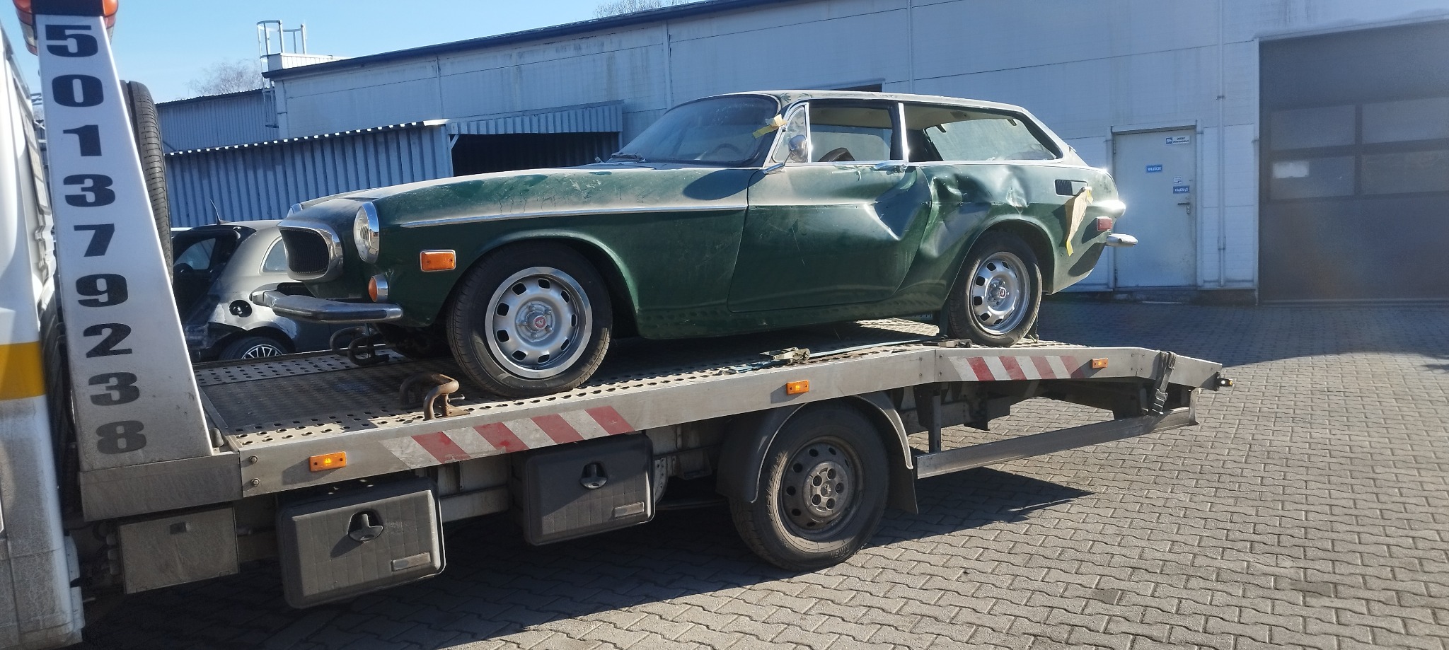 Zielony, uszkodzony Volvo 1800ES na platformie lawety z numerem telefonu, widoczny w tle budynek przemysłowy.