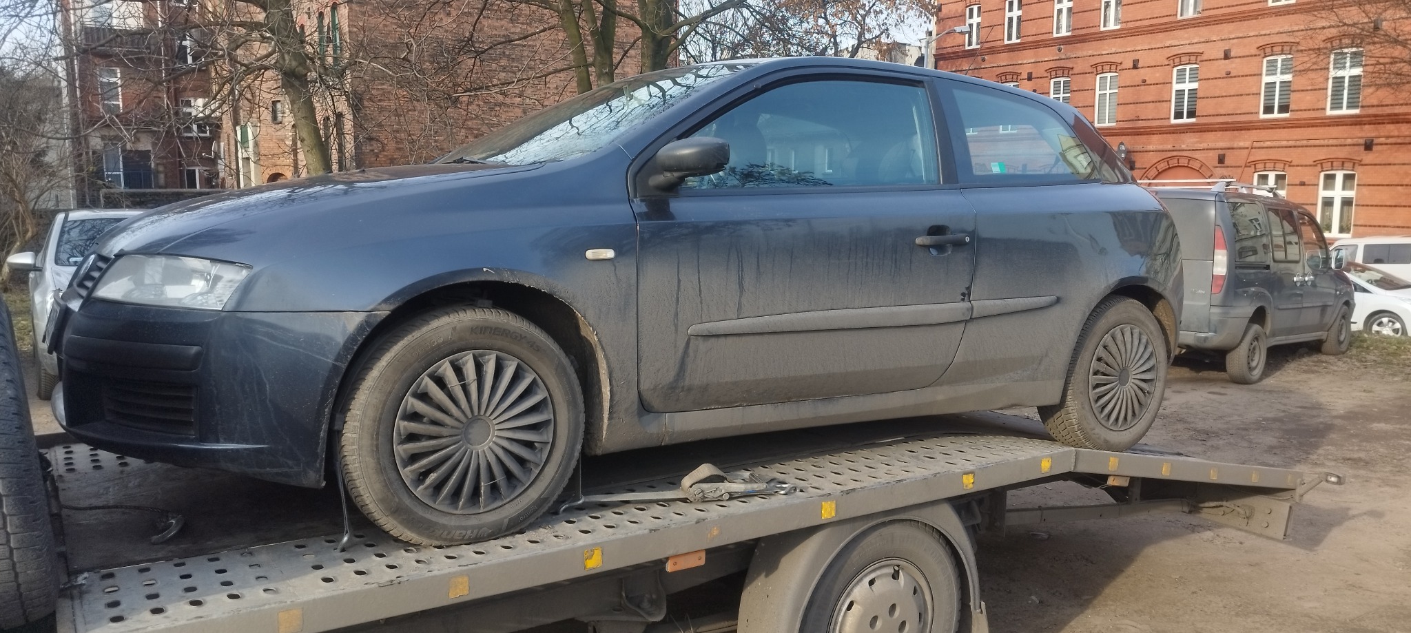 Brudny, granatowy samochód osobowy na platformie lawety, widoczny budynek z czerwonej cegły w tle.