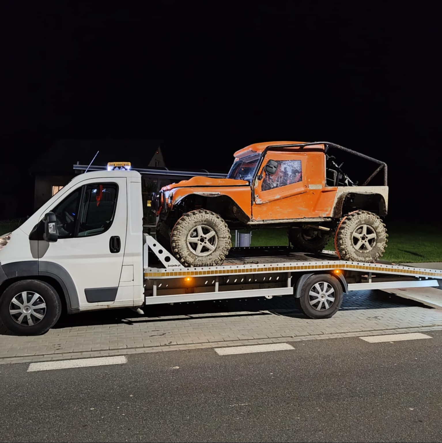 Pomarańczowy, terenowy pojazd na lawecie w nocy. Biała laweta z żółtym światłem ostrzegawczym. Widoczne opony terenowe i konstrukcja klatki bezpieczeństwa pojazdu.