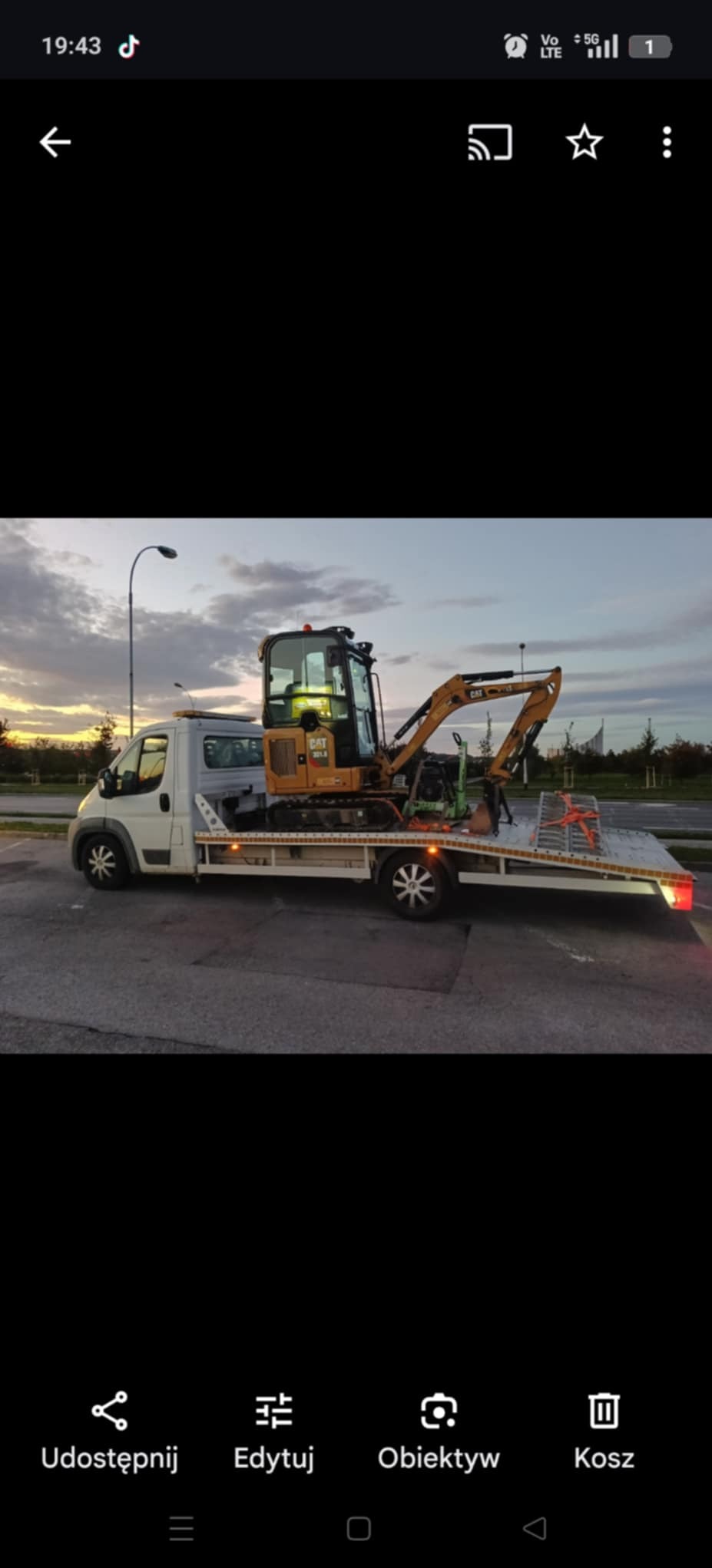 Laweta z pomarańczową mini koparką CAT 301.8 na platformie, widok z boku na tle szarego nieba o zmierzchu. Koła lawety z motywem gwiazdy.