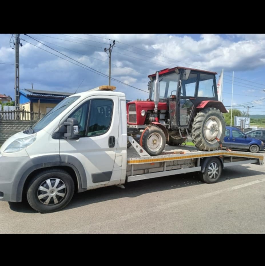 Laweta przewozi czerwony traktor z kabiną na platformie. Biały bus lawety z pomarańczowym światłem ostrzegawczym. Tło: budynki, słupy energetyczne, błękitne niebo.