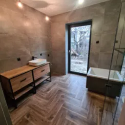 Nowoczesna łazienka z drewnopodobną podłogą ułożoną w jodełkę, szarobrązowe płytki na ścianach, drewniana szafka pod umywalką z czarnymi elementami, wanna przy oknie z widokiem na plac budowy.