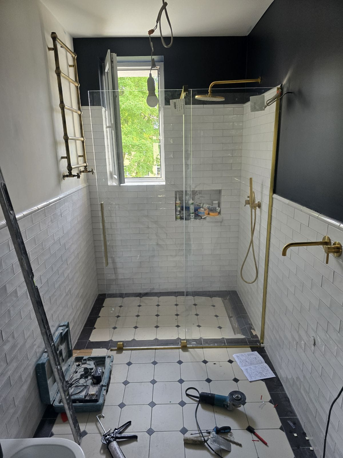 Łazienka w trakcie wykańczania: kabina prysznicowa ze złotymi elementami, białe płytki imitujące cegłę, czarna ściana, narzędzia i skrzynka narzędziowa na podłodze z czarno-białą szachownicą.