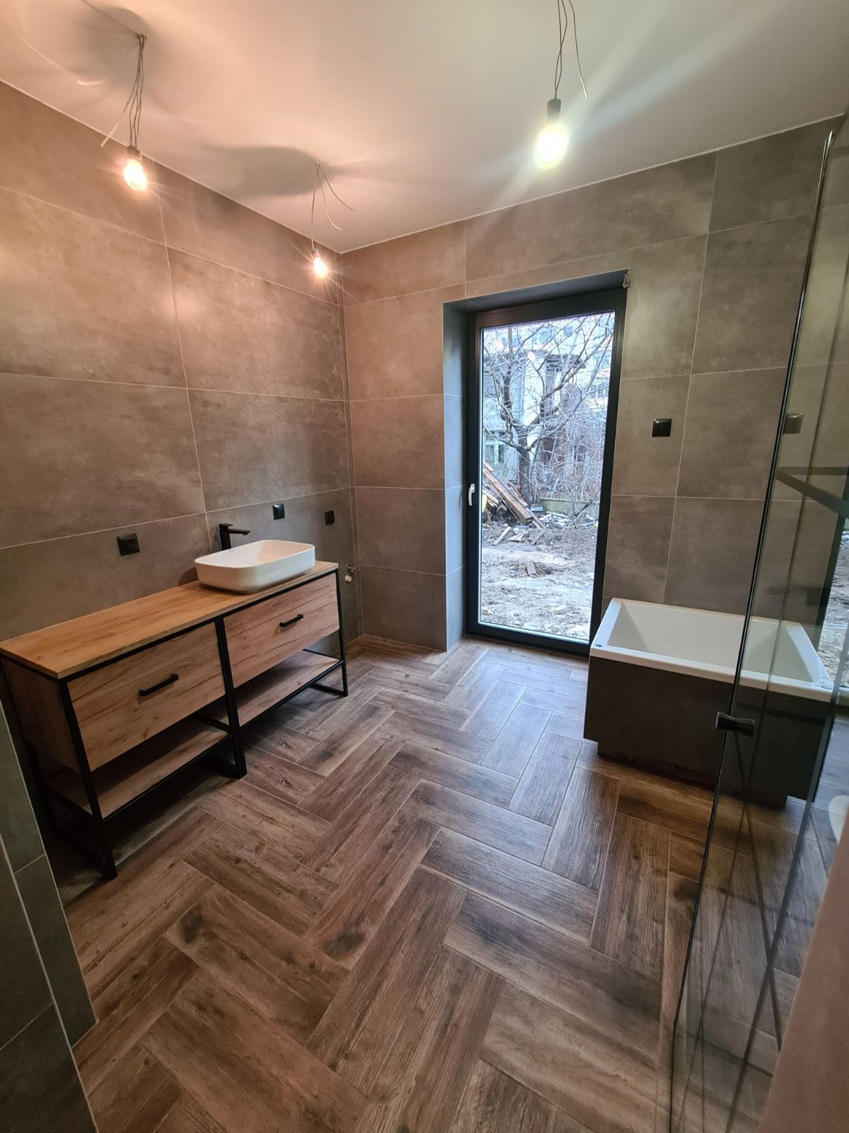 Nowoczesna łazienka z drewnopodobną podłogą ułożoną w jodełkę, szarobrązowe płytki na ścianach, drewniana szafka pod umywalką z czarnymi elementami, wanna przy oknie z widokiem na plac budowy.