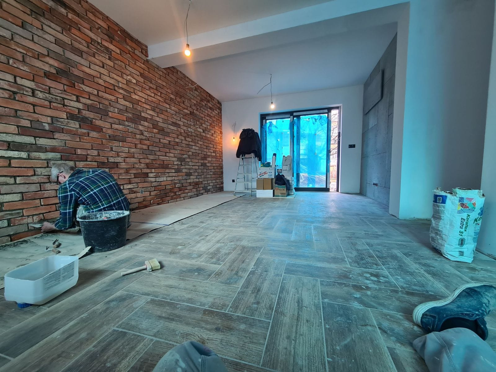 Układanie cegieł na ścianie w przestronnym wnętrzu, widoczne narzędzia, wiadro z zaprawą oraz ułożona podłoga w jodełkę, w tle okno zasłonięte folią.