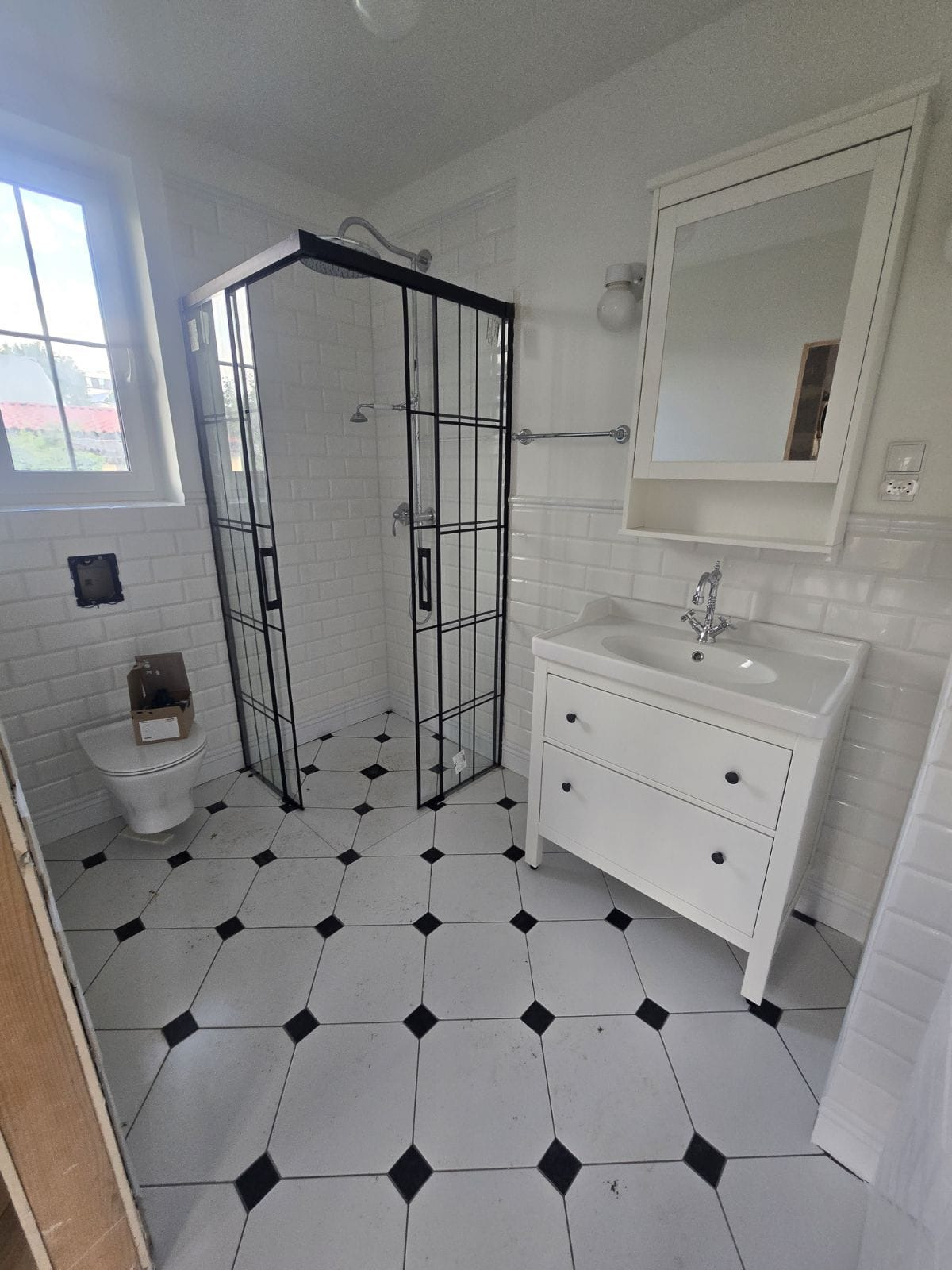 Biała łazienka z kabiną prysznicową typu walk-in z czarnymi ramami, białą umywalką z szafką i biało-czarną podłogą w romby, widoczna toaleta z kartonem.
