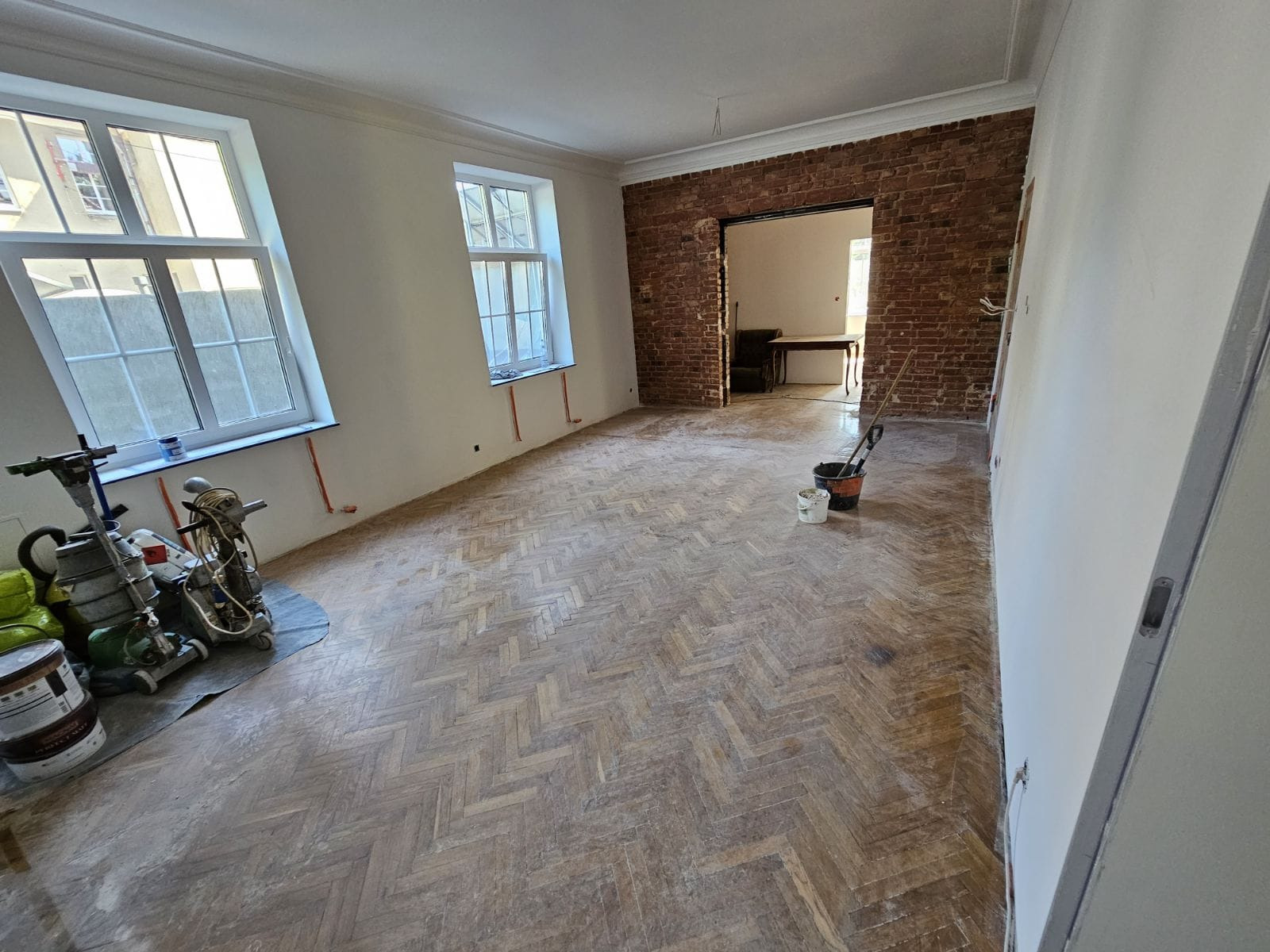 Pokój w trakcie renowacji z parkietem ułożonym we wzór jodełki, szlifierka do parkietu, odsłonięta ceglana ściana i przygotowane farby.