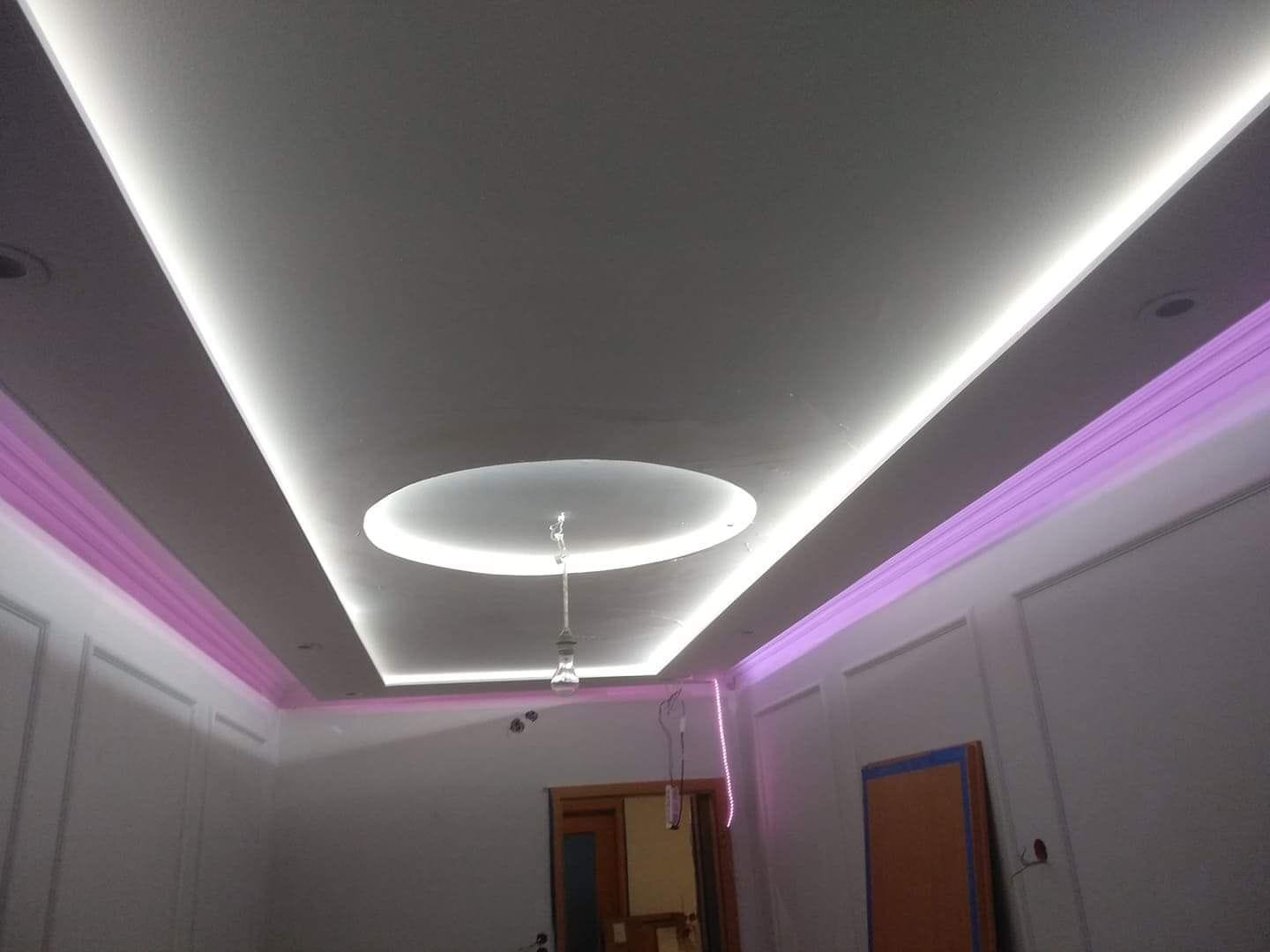 Nowoczesny sufit podwieszany z oświetleniem LED: białe światło wzdłuż krawędzi i okrągły element centralny, uzupełniony fioletowym oświetleniem nad ścianami.