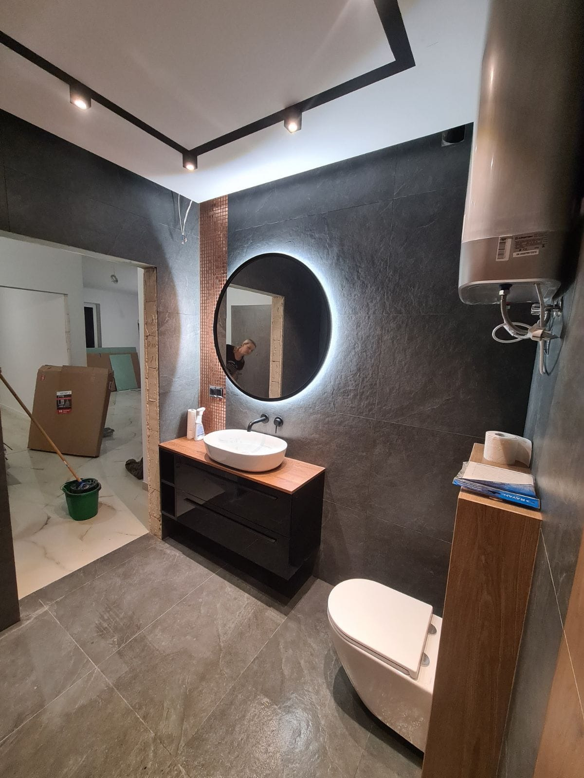 Łazienka w trakcie wykańczania z czarnymi płytkami, okrągłym lustrem z podświetleniem LED, białą umywalką nablatową i toaletą, widoczne kartony i wiadro w przejściu do innego pomieszczenia.