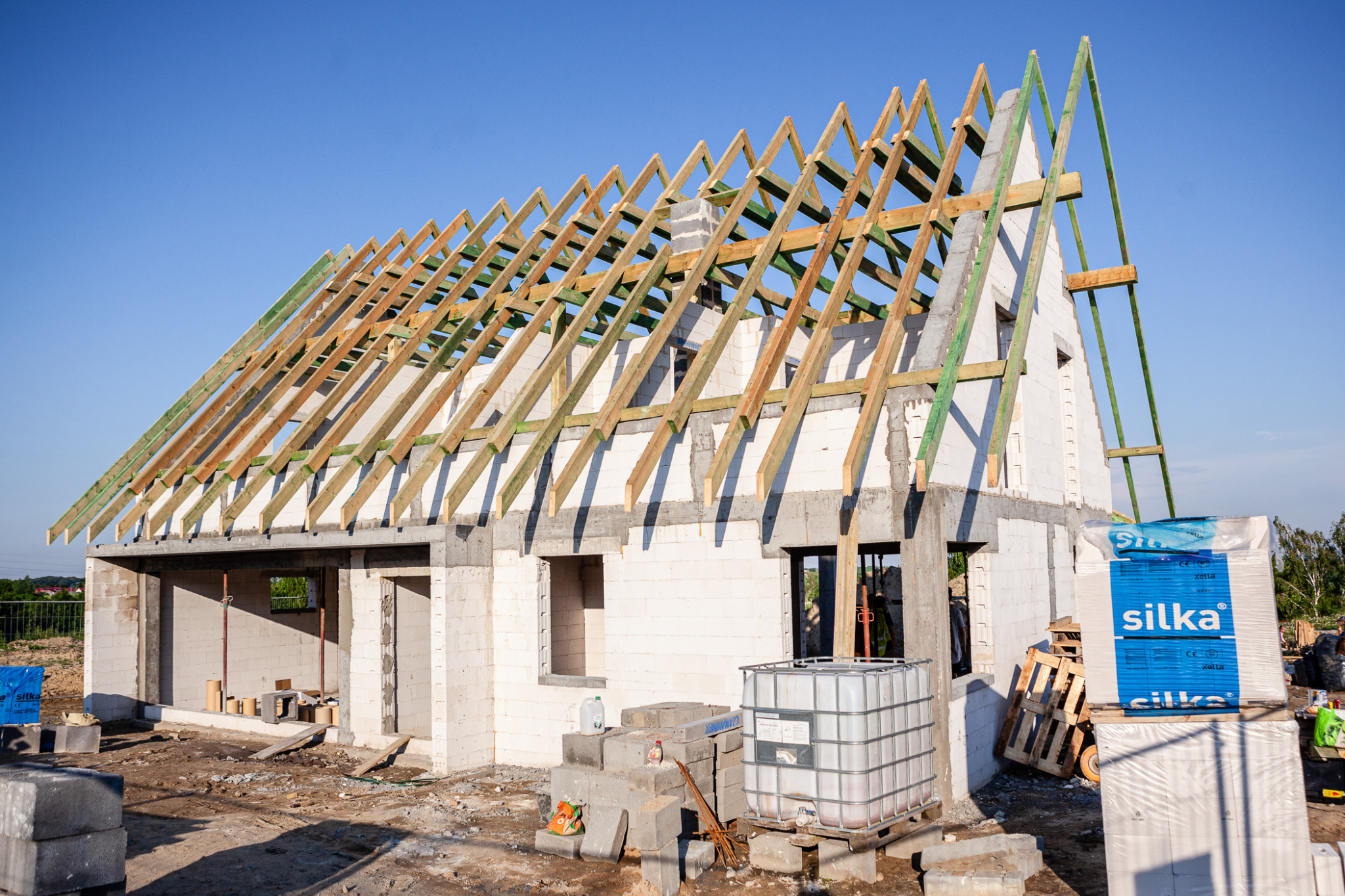 Surowy stan domu z białych bloczków z widoczną konstrukcją drewnianą dachu, paletami z materiałami Silka i zbiornikiem IBC na placu budowy w słoneczny dzień.