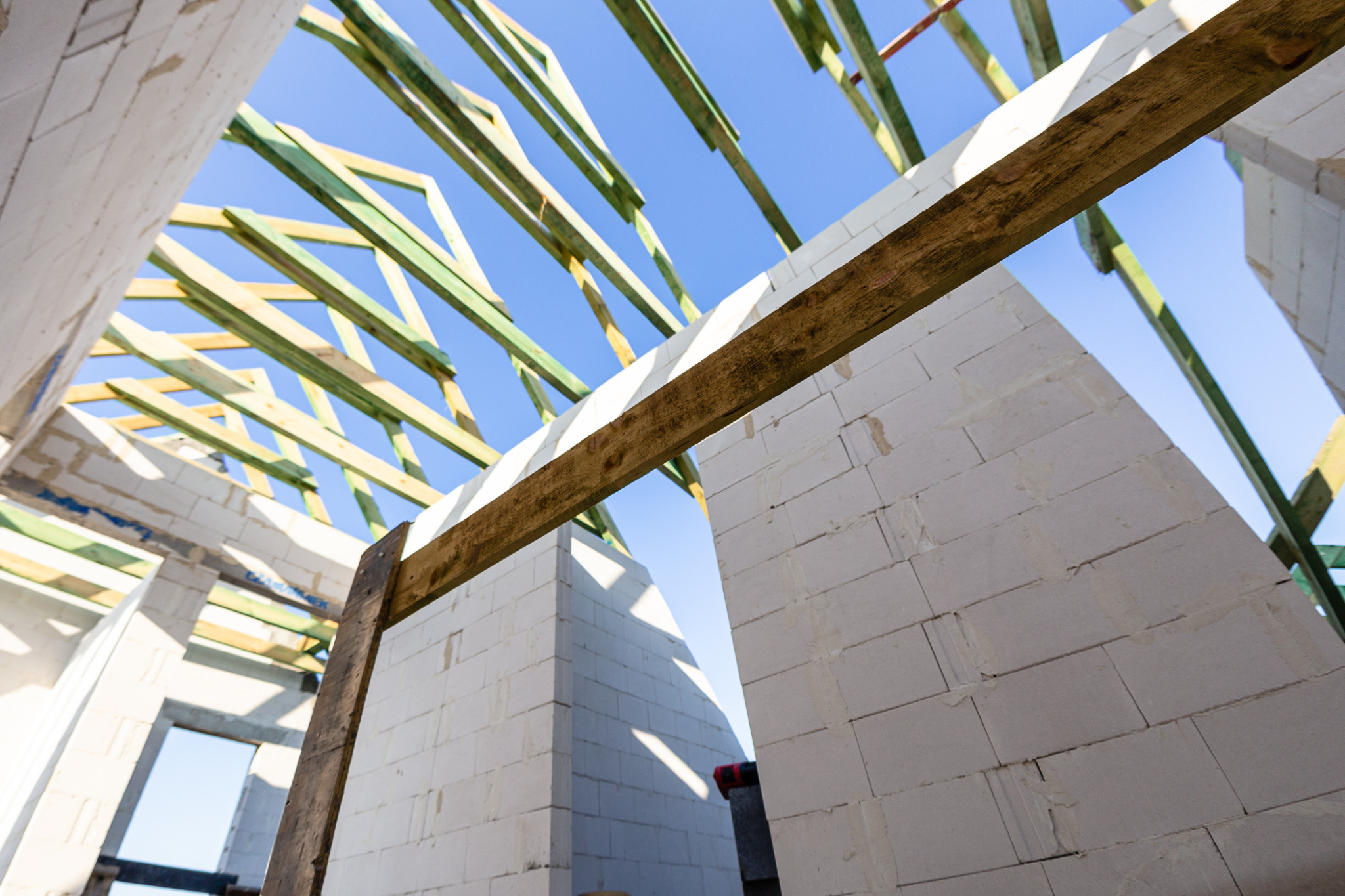 Wnętrze domu w trakcie budowy, ściany z białych bloczków, drewniane elementy konstrukcji dachu na tle błękitnego nieba.