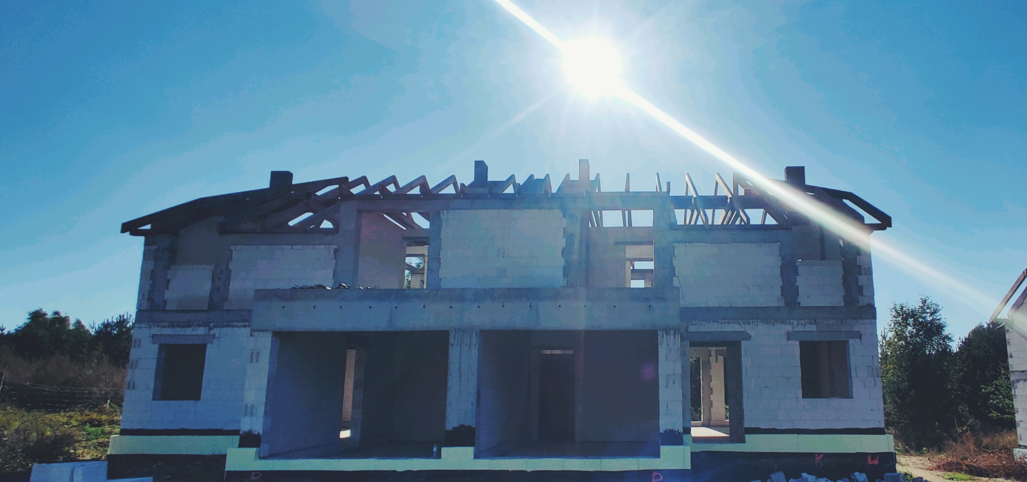 Surowy stan dwukondygnacyjnego domu z pustaków, z widoczną konstrukcją dachu i niezadaszonymi otworami okiennymi, otoczony zielenią w słoneczny dzień.