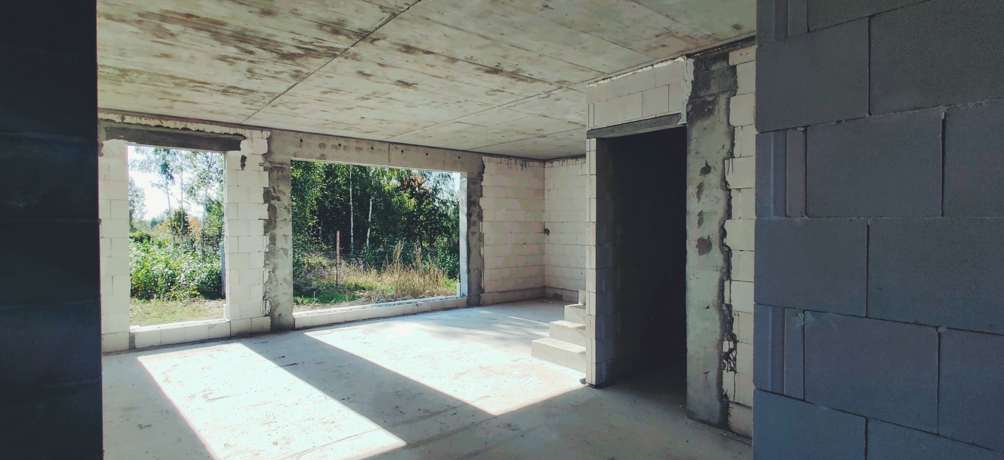 Wnętrze domu w stanie surowym otwartym z widocznymi ścianami z bloczków, betonowym stropem i oknami wychodzącymi na zieleń.