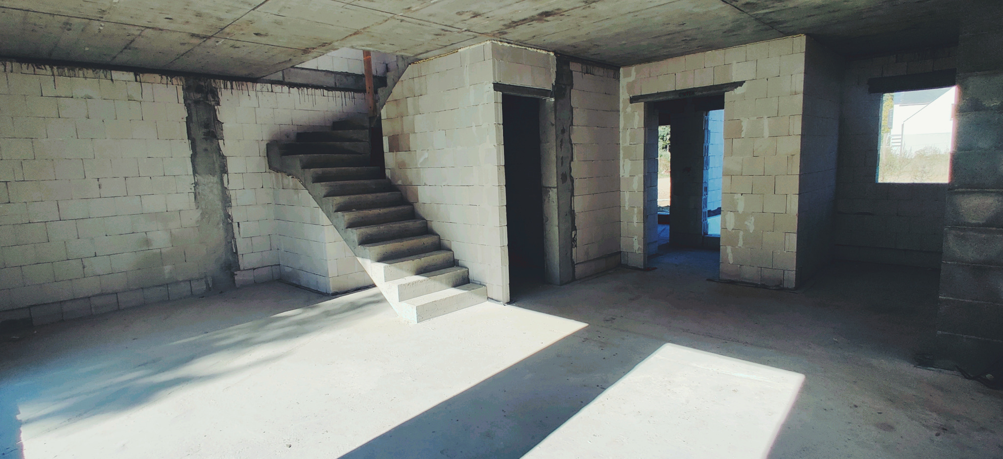 Wnętrze surowego domu w trakcie budowy, ściany z białych bloczków, betonowe schody, widoczne otwory na drzwi i okno, surowy betonowy strop.