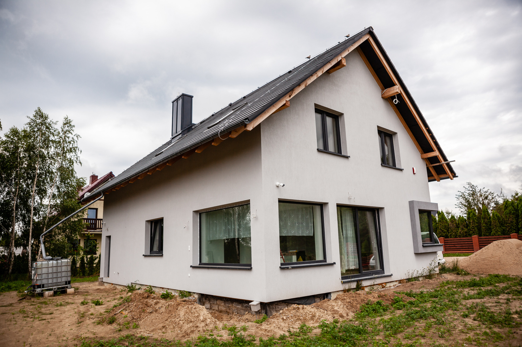 Nowoczesny, jasnoszary dom jednorodzinny z czarną dachówką i oknami, widoczny z zewnątrz, z fragmentem ogrodu, zbiornikiem na wodę deszczową i stertą piasku obok