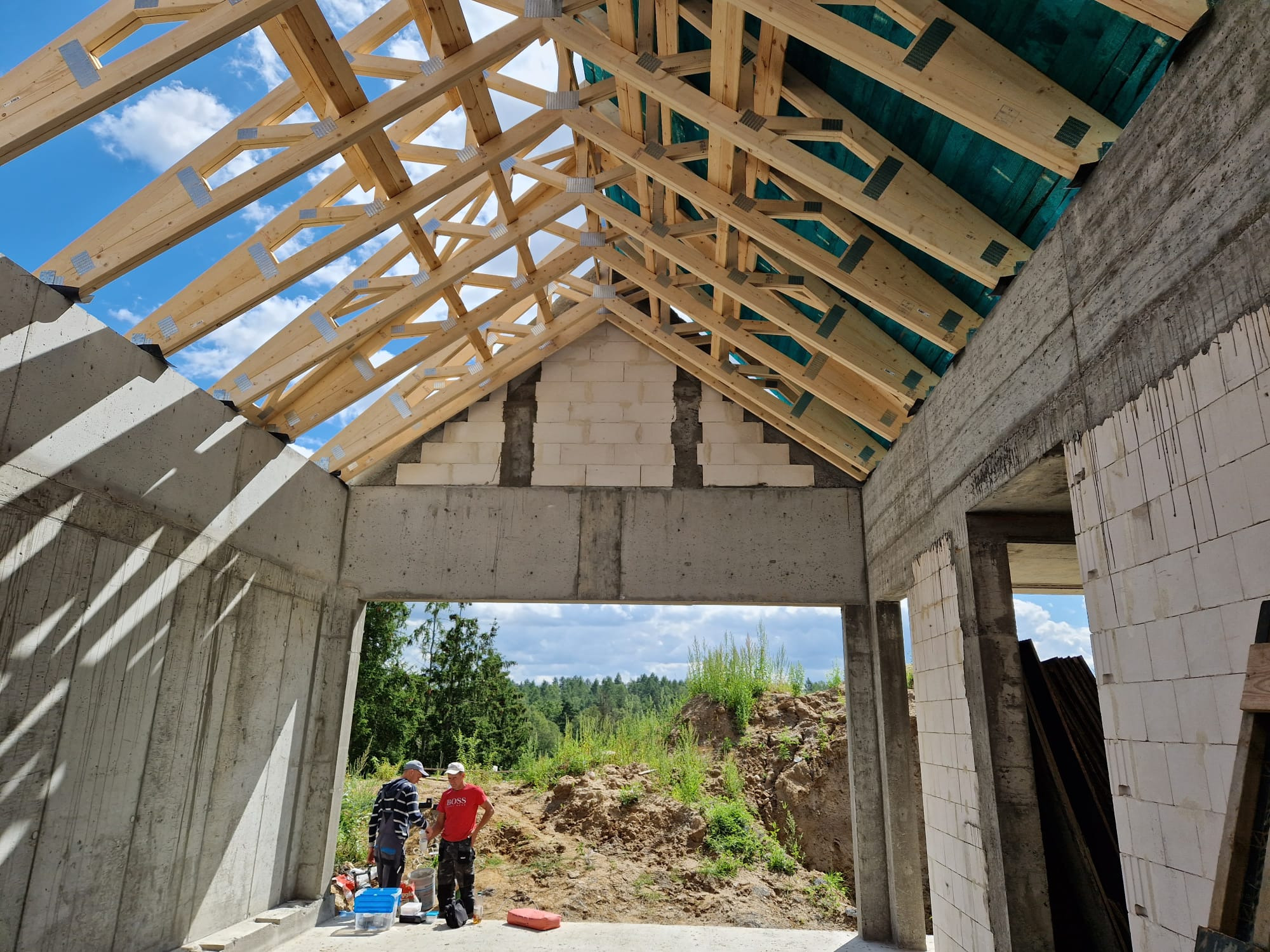 Wnętrze surowego domu w trakcie budowy, widoczne betonowe ściany, strop z pustaków, drewniana konstrukcja dachu z membraną, dwóch pracowników na zewnątrz budynku