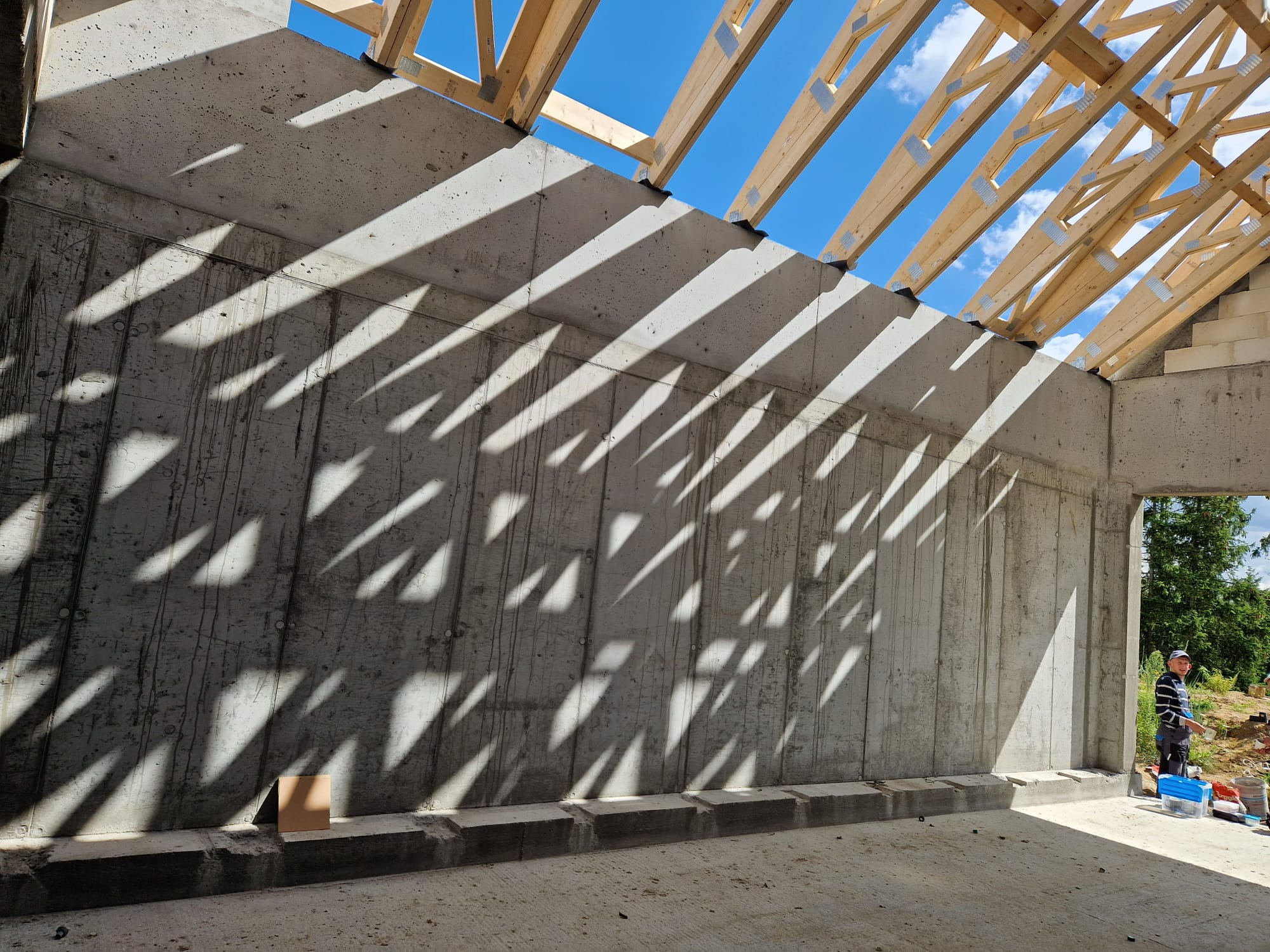 Surowa betonowa ściana w budowie domu z widocznymi cieniami drewnianej konstrukcji dachu, z pracownikiem w tle i narzędziami.