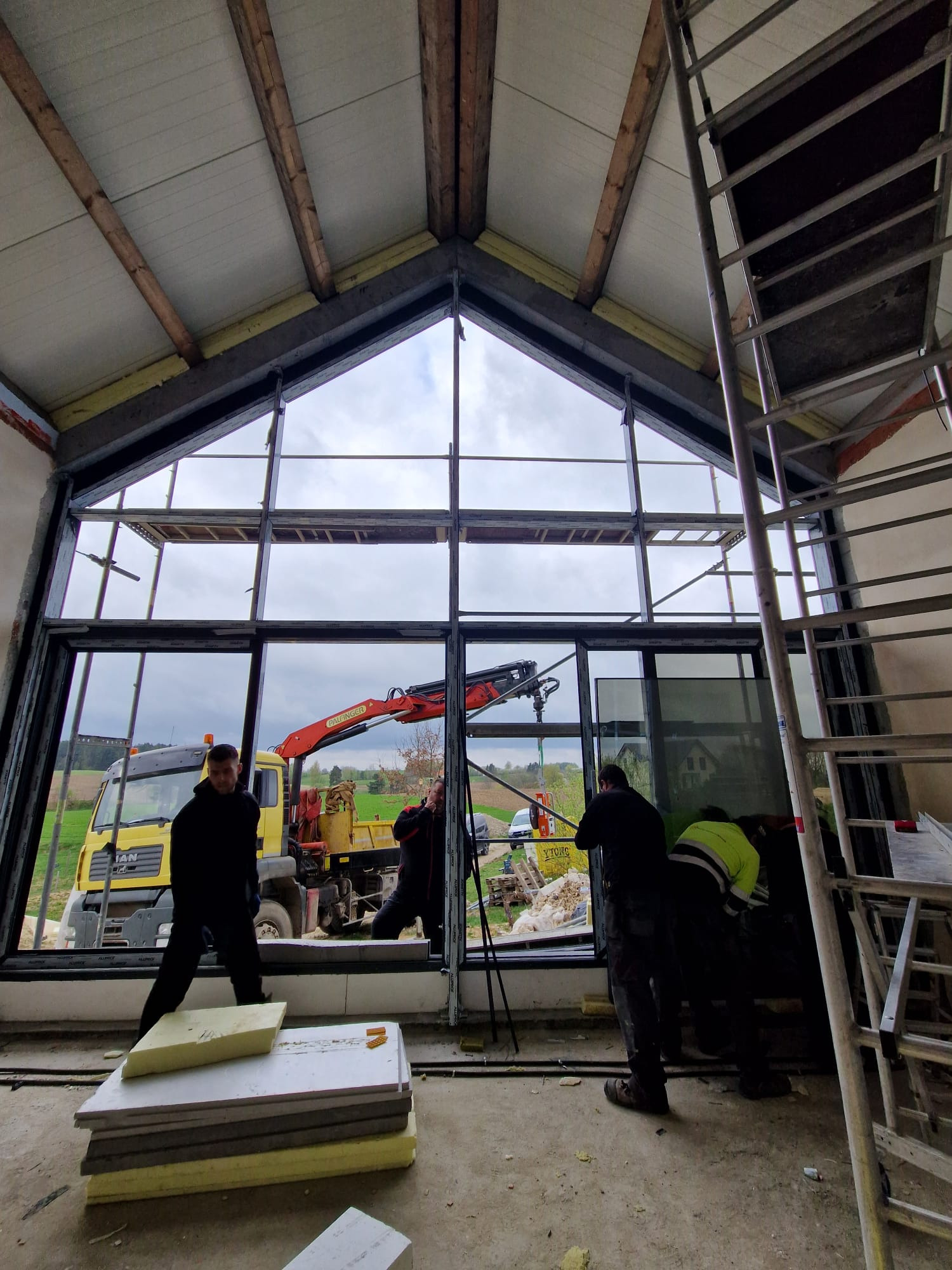 Montaż okna panoramicznego w surowym stanie budynku, z użyciem dźwigu samochodowego; widoczne płyty izolacyjne, rusztowanie i pracownicy w trakcie instalacji.