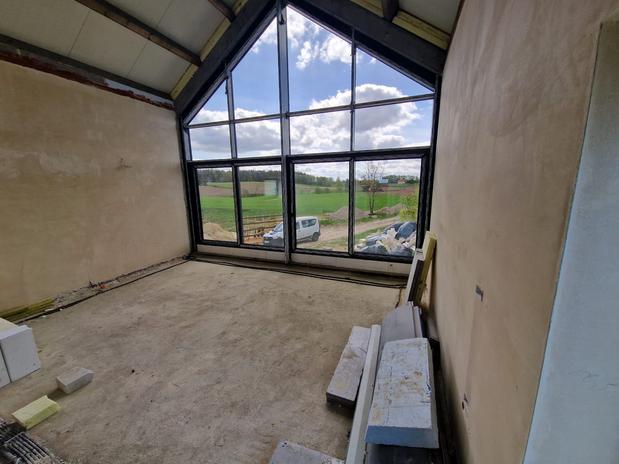Wnętrze w stanie surowym z dużym oknem szklanym z widokiem na krajobraz, widoczny samochód dostawczy i materiały budowlane.