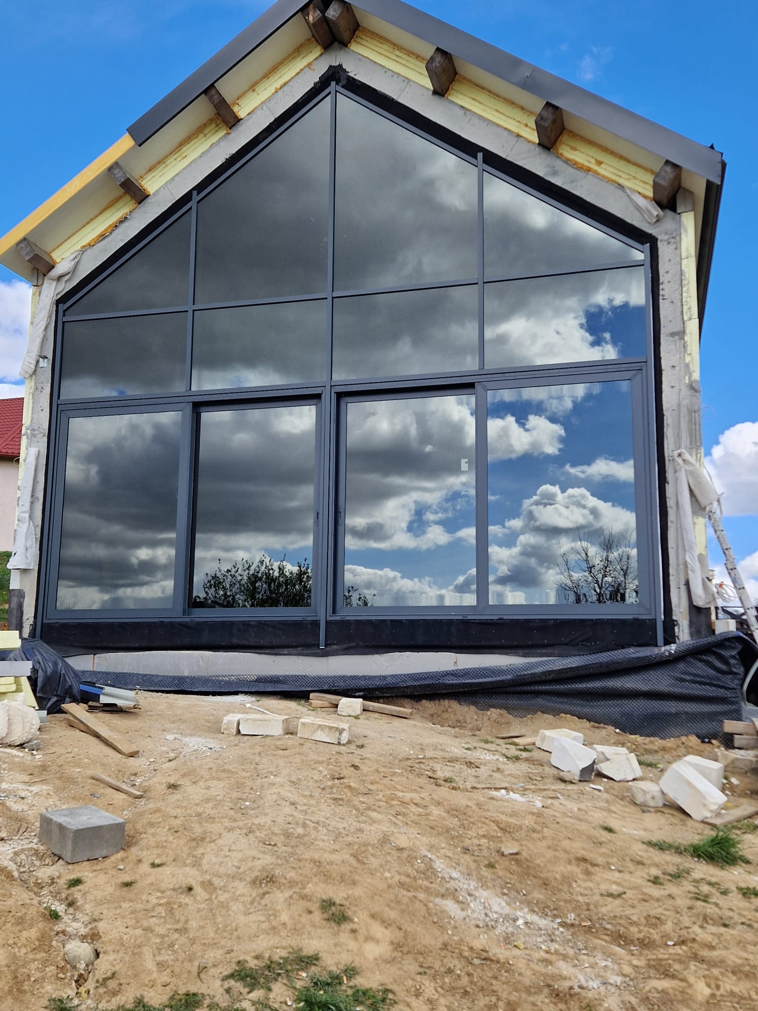 Surowy stan domu murowanego z dużym oknem z czarną ramą, w którym odbijają się chmury i drzewa, widoczne materiały izolacyjne i porozrzucane bloczki betonowe.