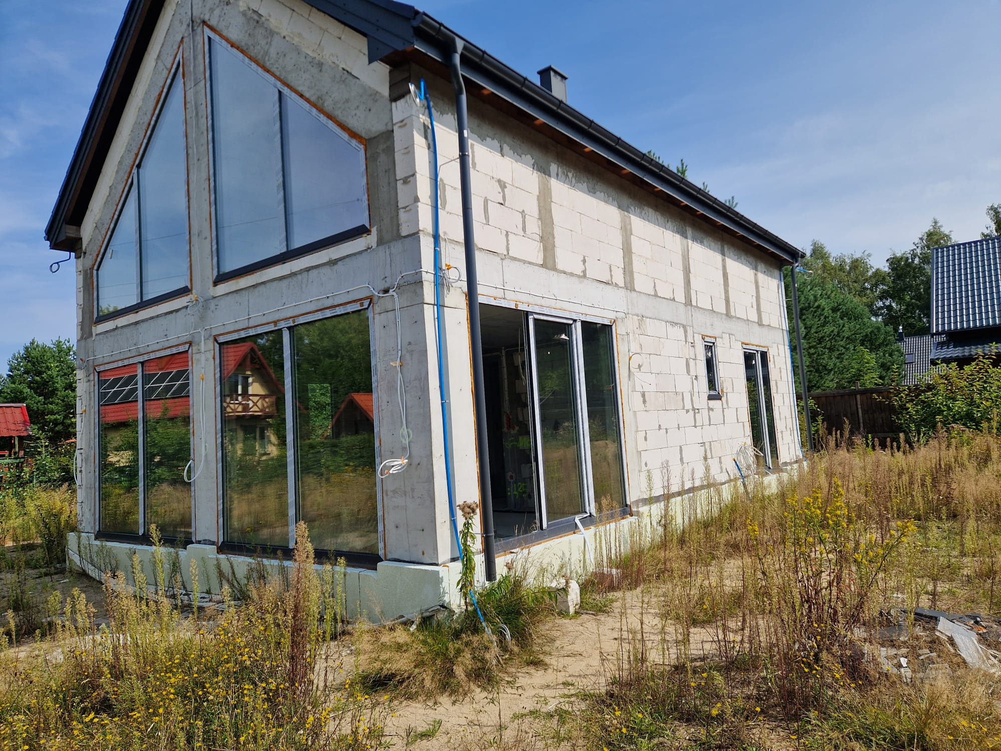 Surowy stan domu z betonu komórkowego z dużymi oknami, widoczne instalacje na elewacji, otoczenie zarośnięte trawą i chwastami.