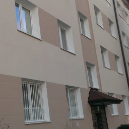 Miwobud - Wielopiętrowy budynek z elewacją w odcieniach beżu, z białymi ramami okiennymi i kratami w oknach na parterze, widoczny z perspektywy lekko od dołu.